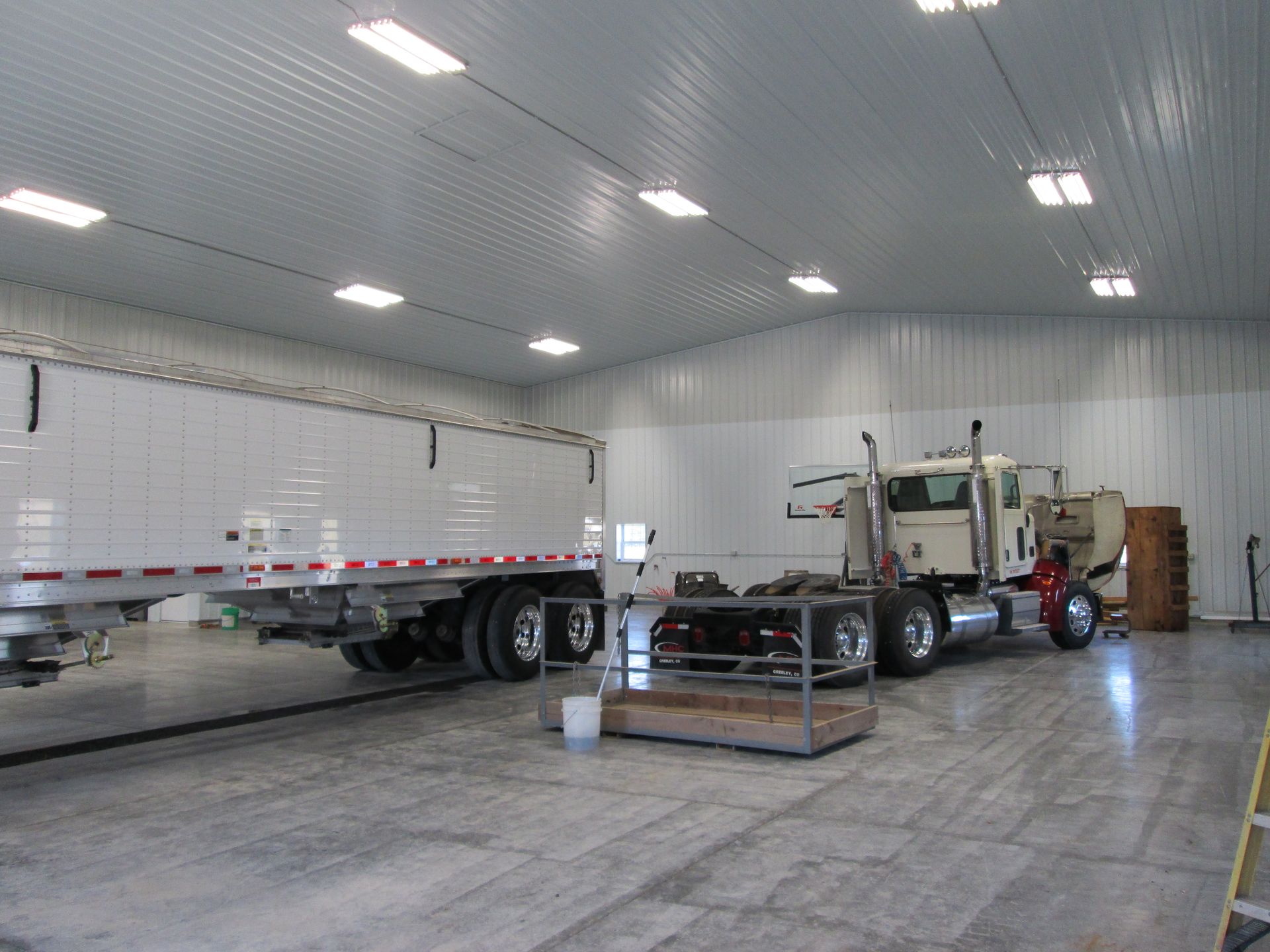 Semi-truck and tractor parked inside a bright metal workshop with a concrete floor.