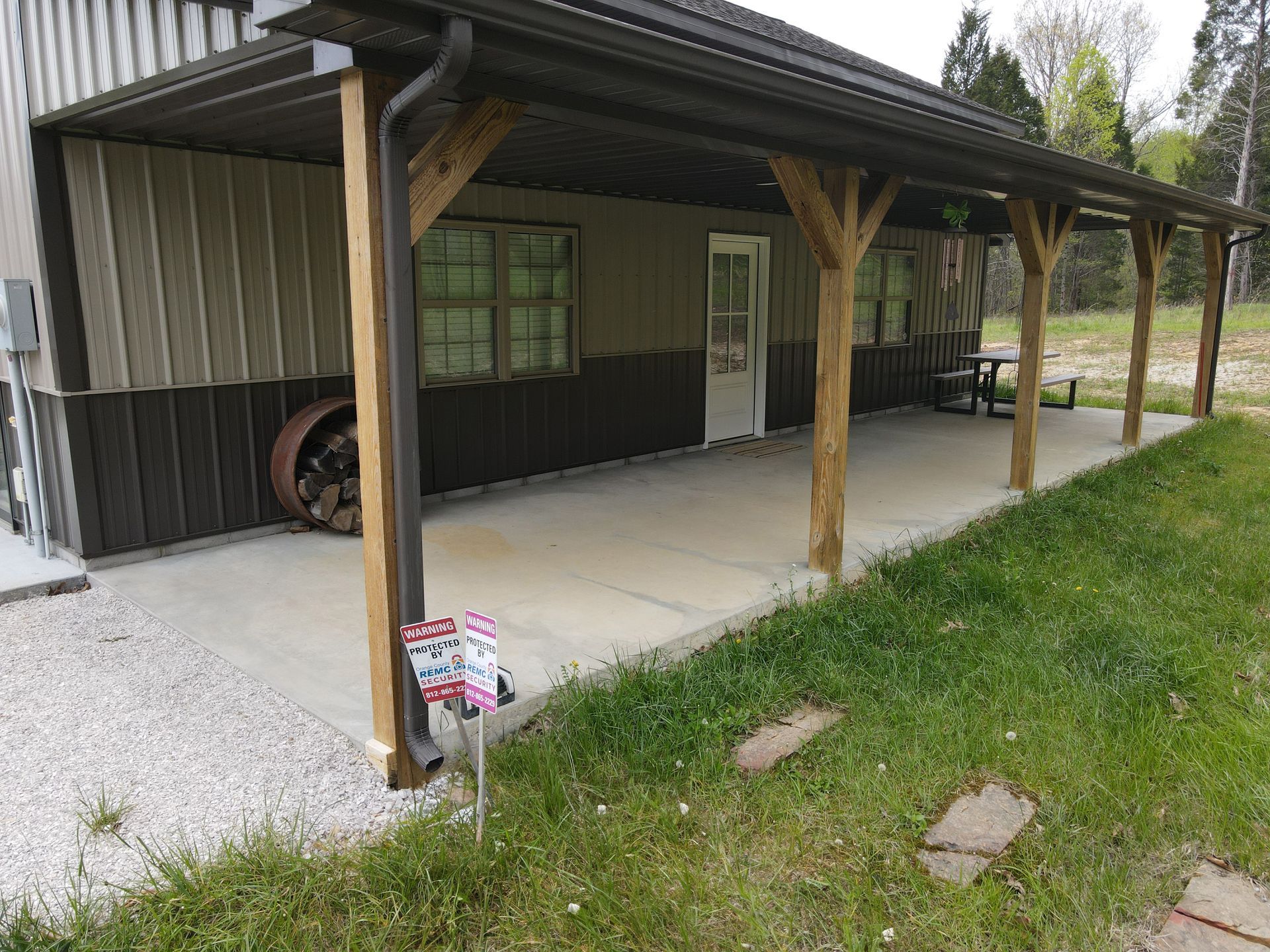 Covered porch with wooden posts beside a dark green house, gravel driveway, and grassy yard.
