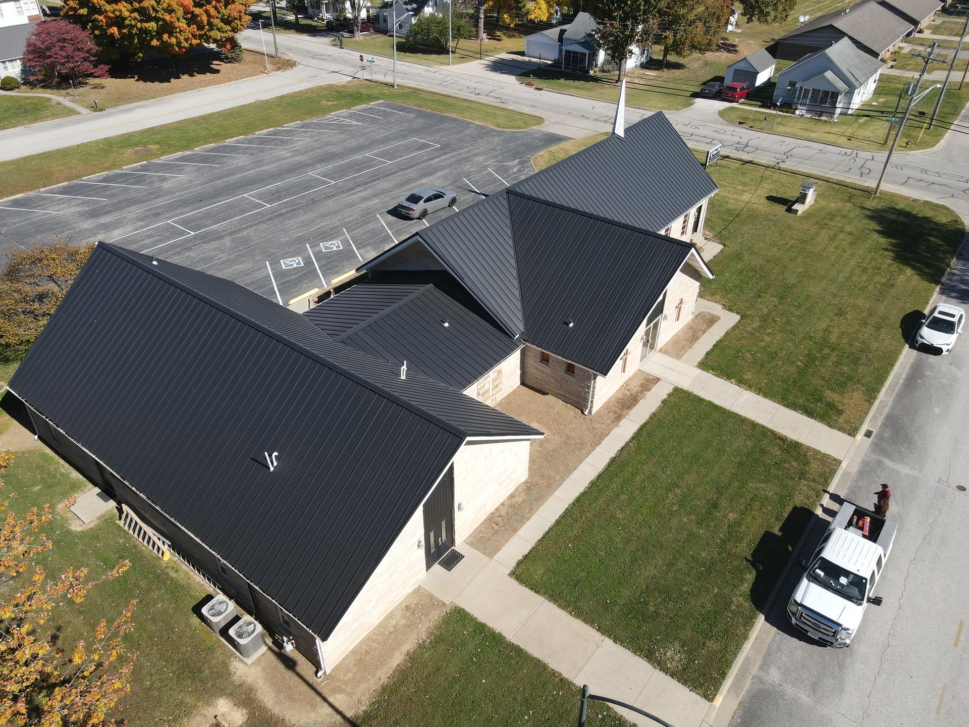 Aerial view of a beige building with dark roofs beside a parking lot and grassy paths.