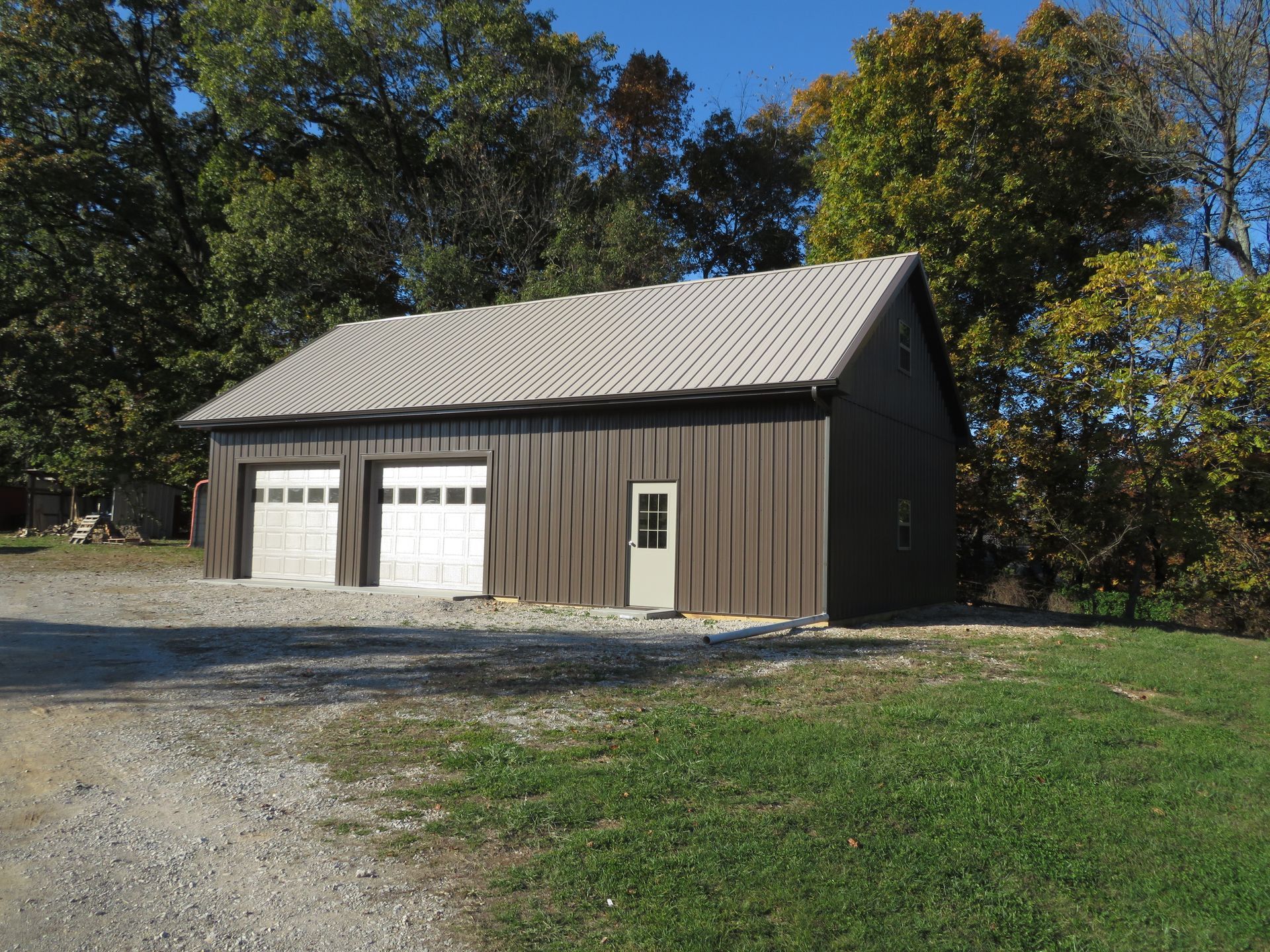 Brown barn with white garage doors and a small side door, beside a gravel drive and grassy yard under autumn trees