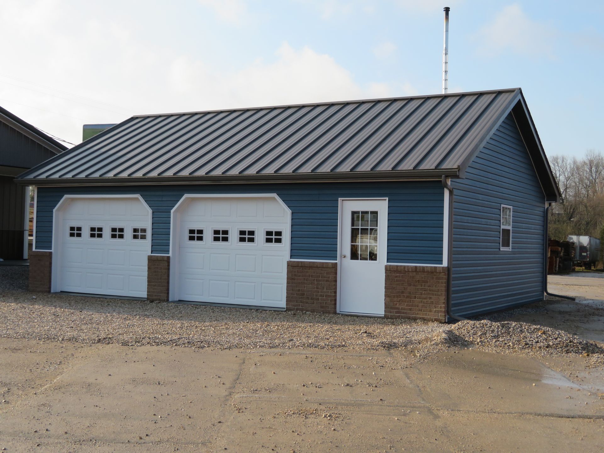 Blue two-car garage with metal roof and white doors on a gravel lot