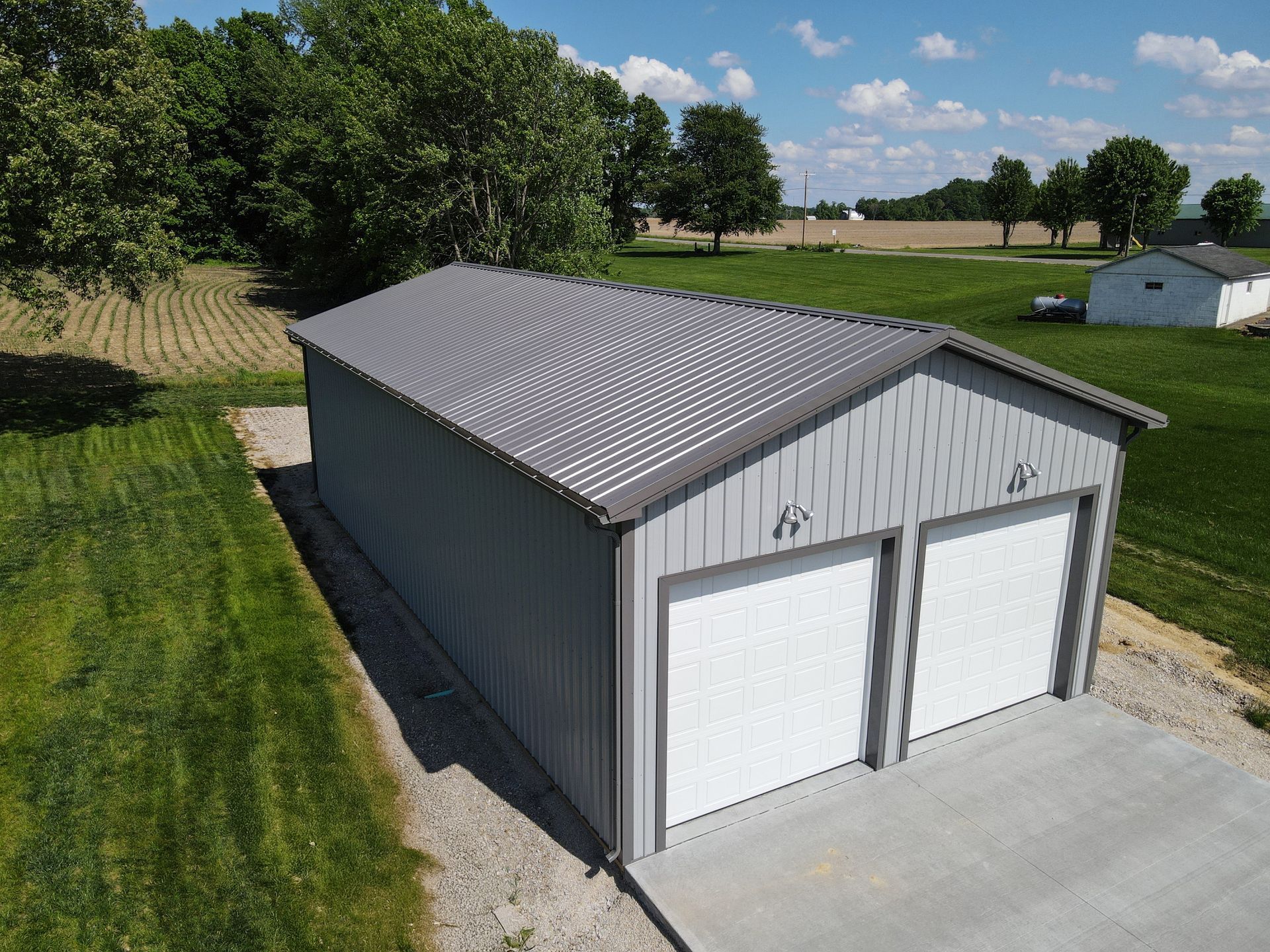 Gray metal two-car garage with white doors on a concrete driveway beside a grassy field under a blue sky