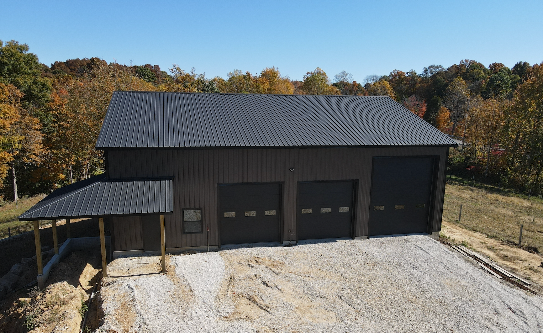 Large dark metal workshop with three garage doors and a covered side porch on a gravel driveway.