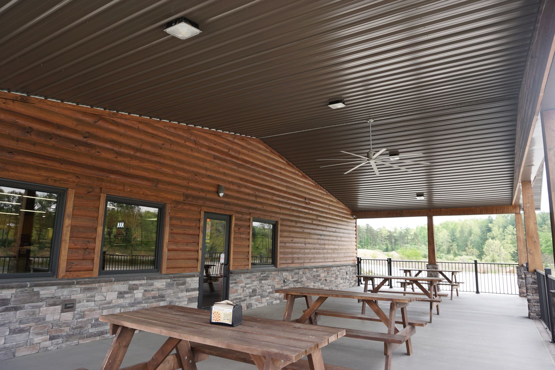 Covered outdoor patio with wooden picnic tables, brick wall, and stone accents beside a wooded view