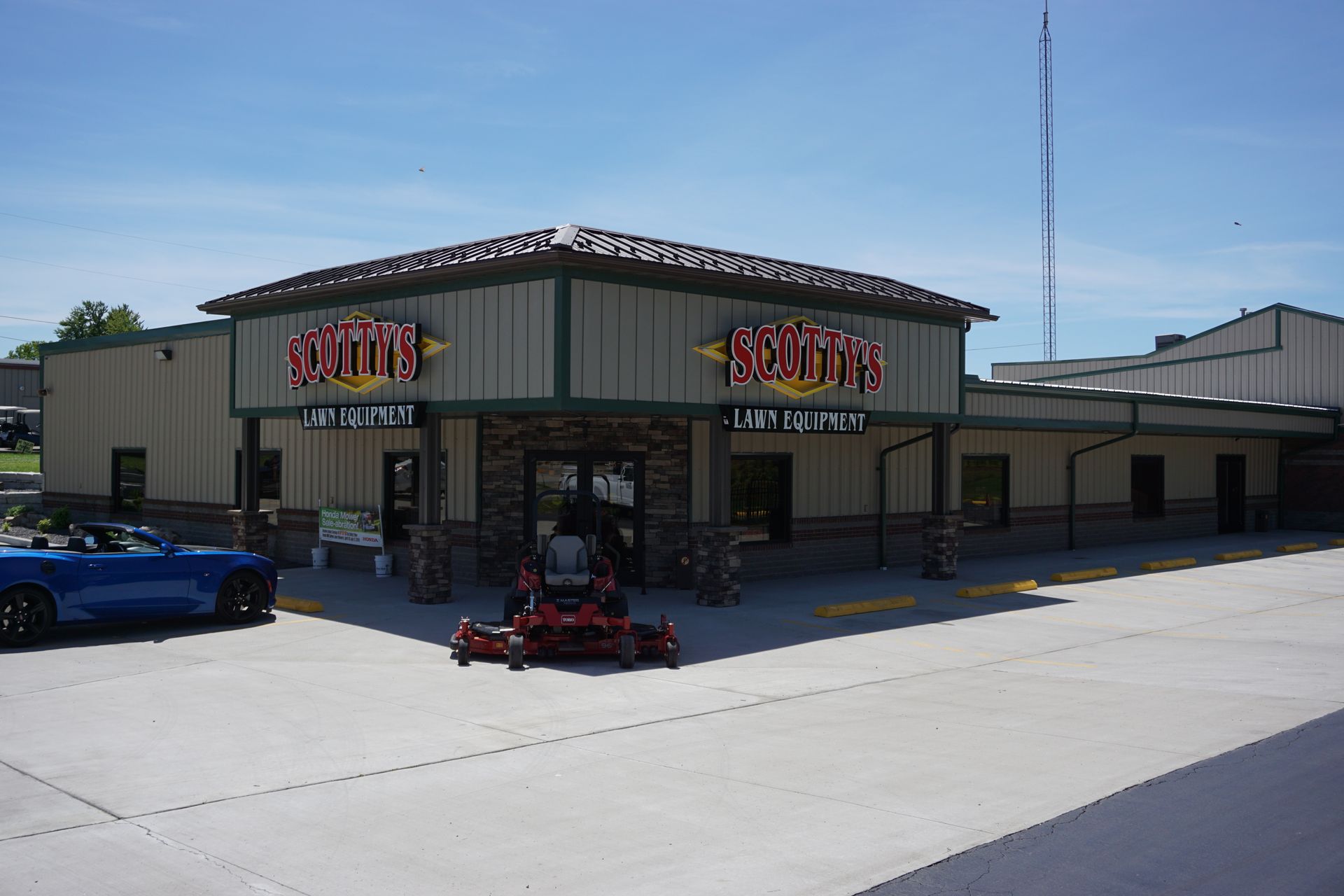 Storefront with colorful signs, red lawn mower display, and blue car in a sunny parking lot.