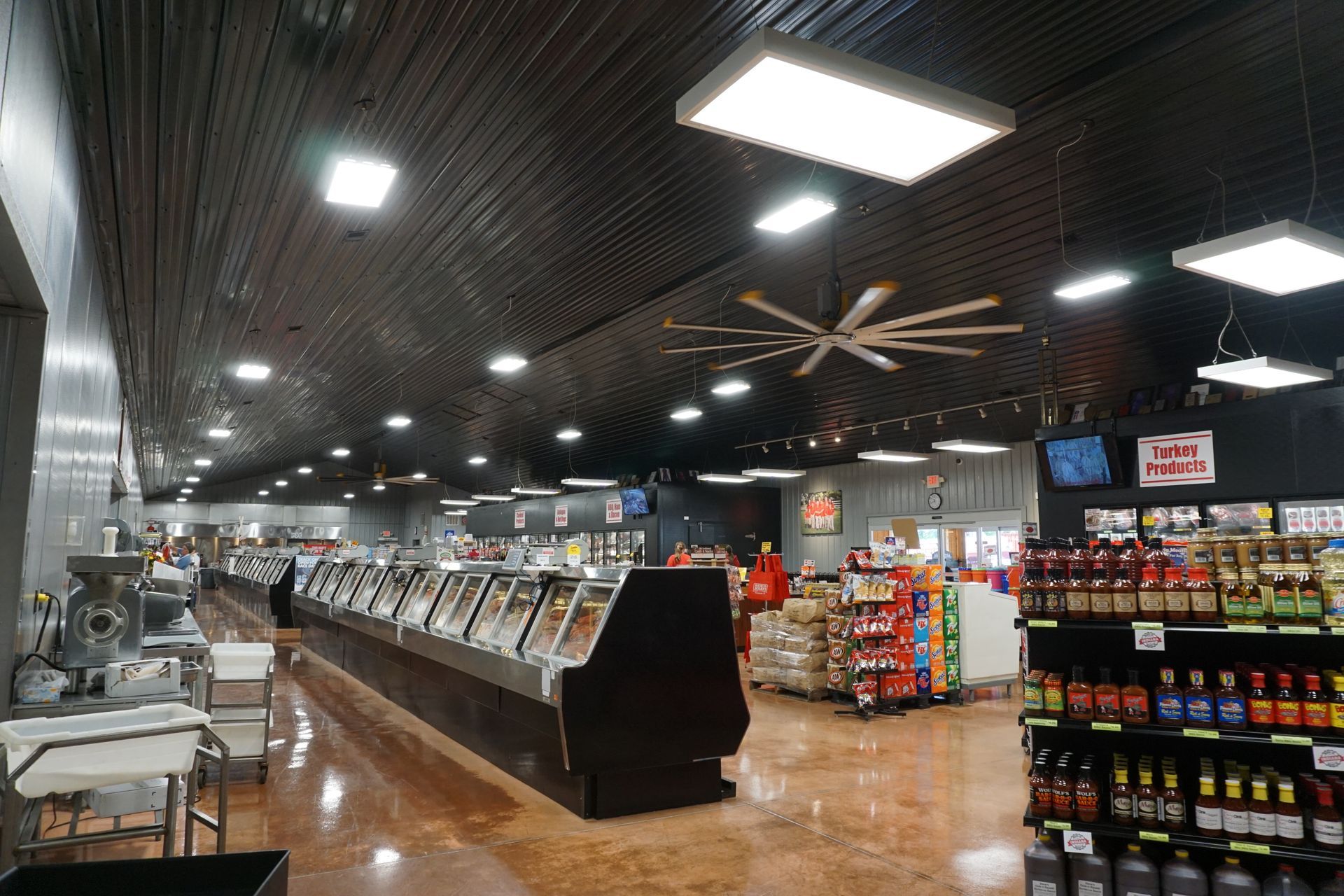 Grocery store deli aisle with refrigerated cases, shelves of snacks, and bright ceiling lights