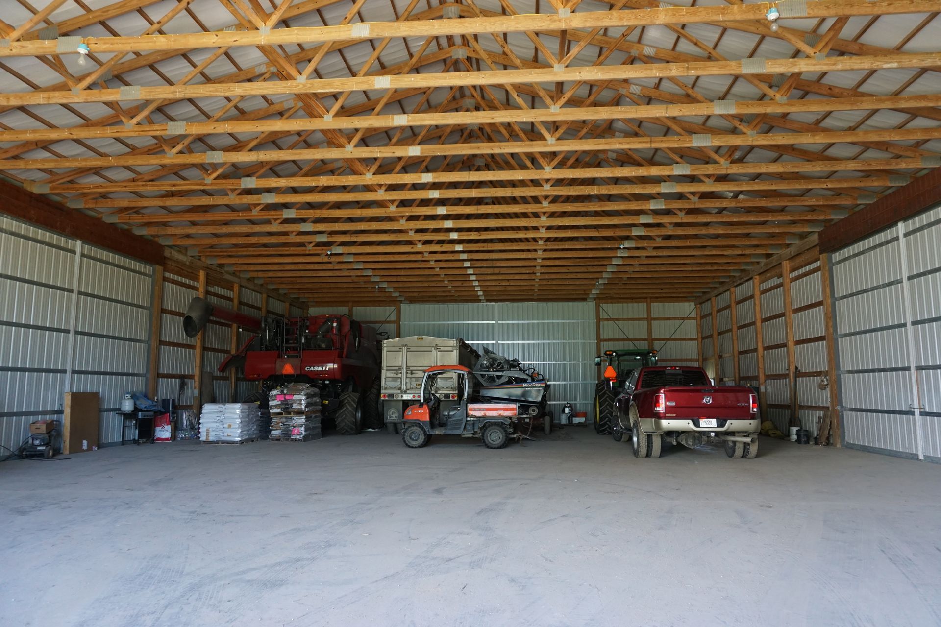 Large open garage with exposed wooden trusses, a tractor, a red pickup, and storage shelves inside.