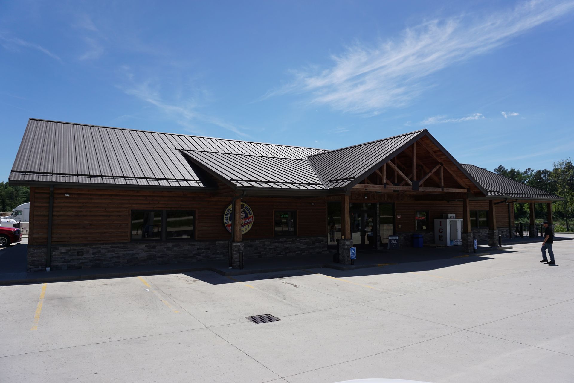 Log cabin-style building with a large wooden roof under a bright blue sky, two people nearby