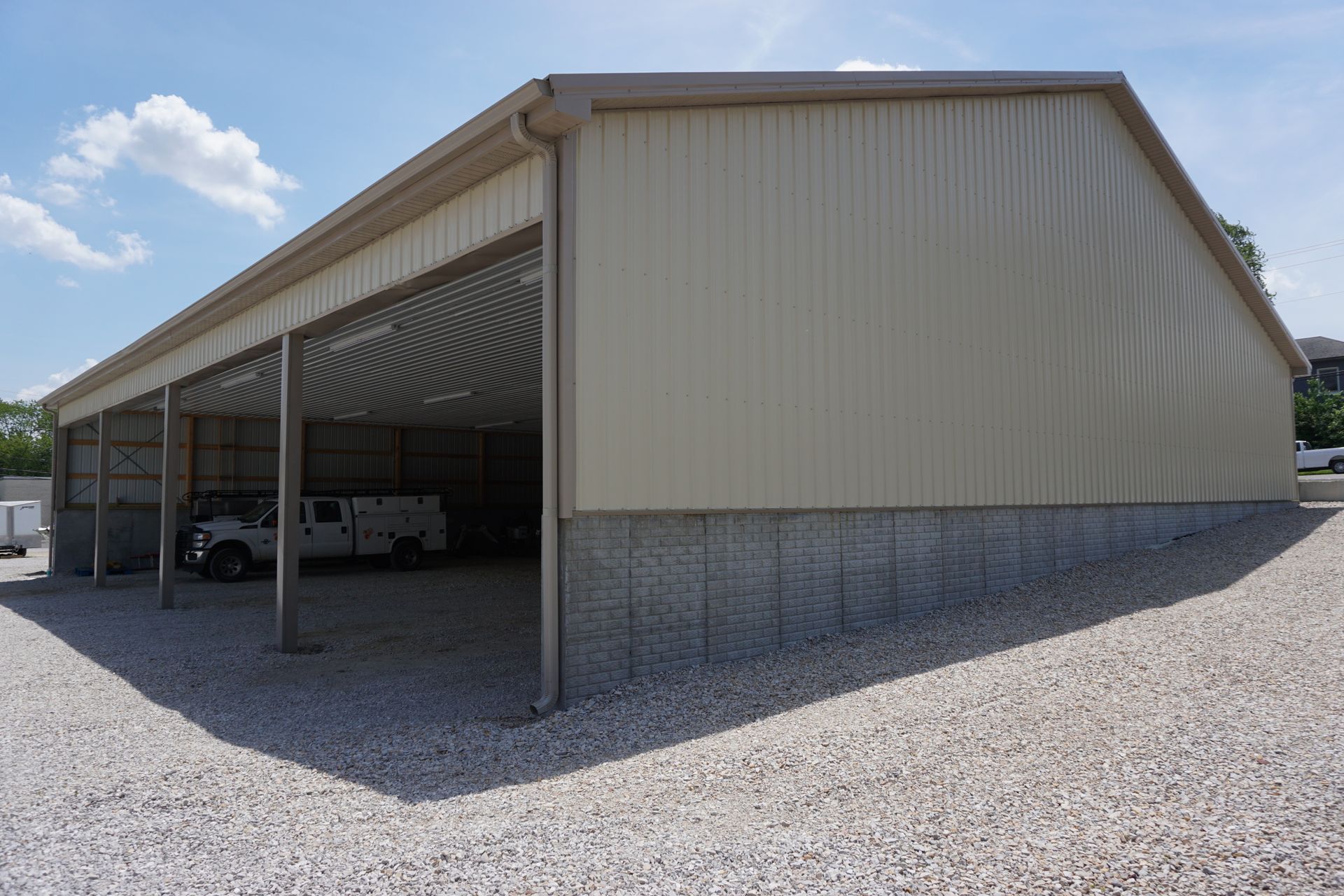 Large metal storage shed with open bays and parked vehicles on a gravel lot under a blue sky