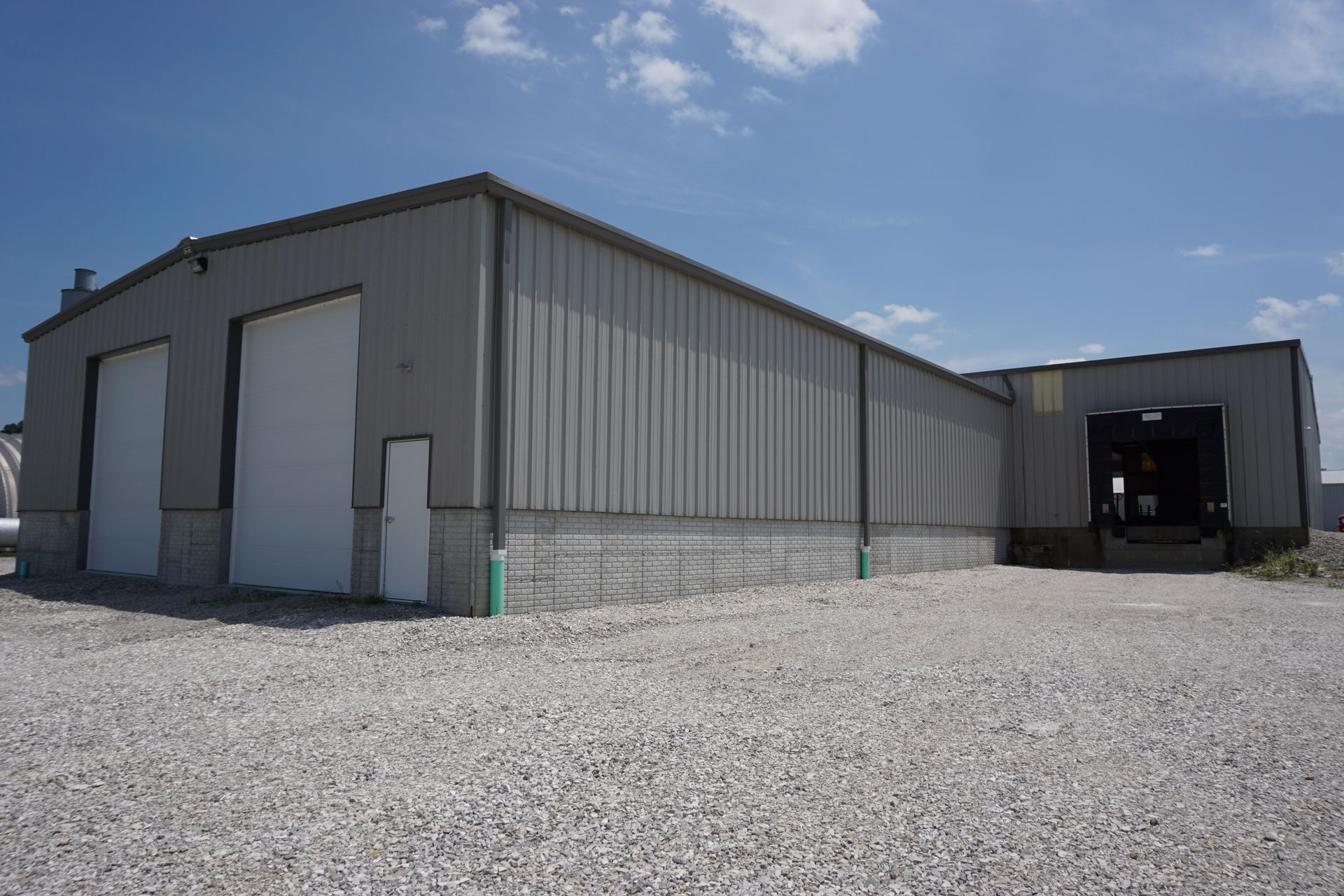 Gray industrial warehouse with large garage doors and gravel lot under a blue sky