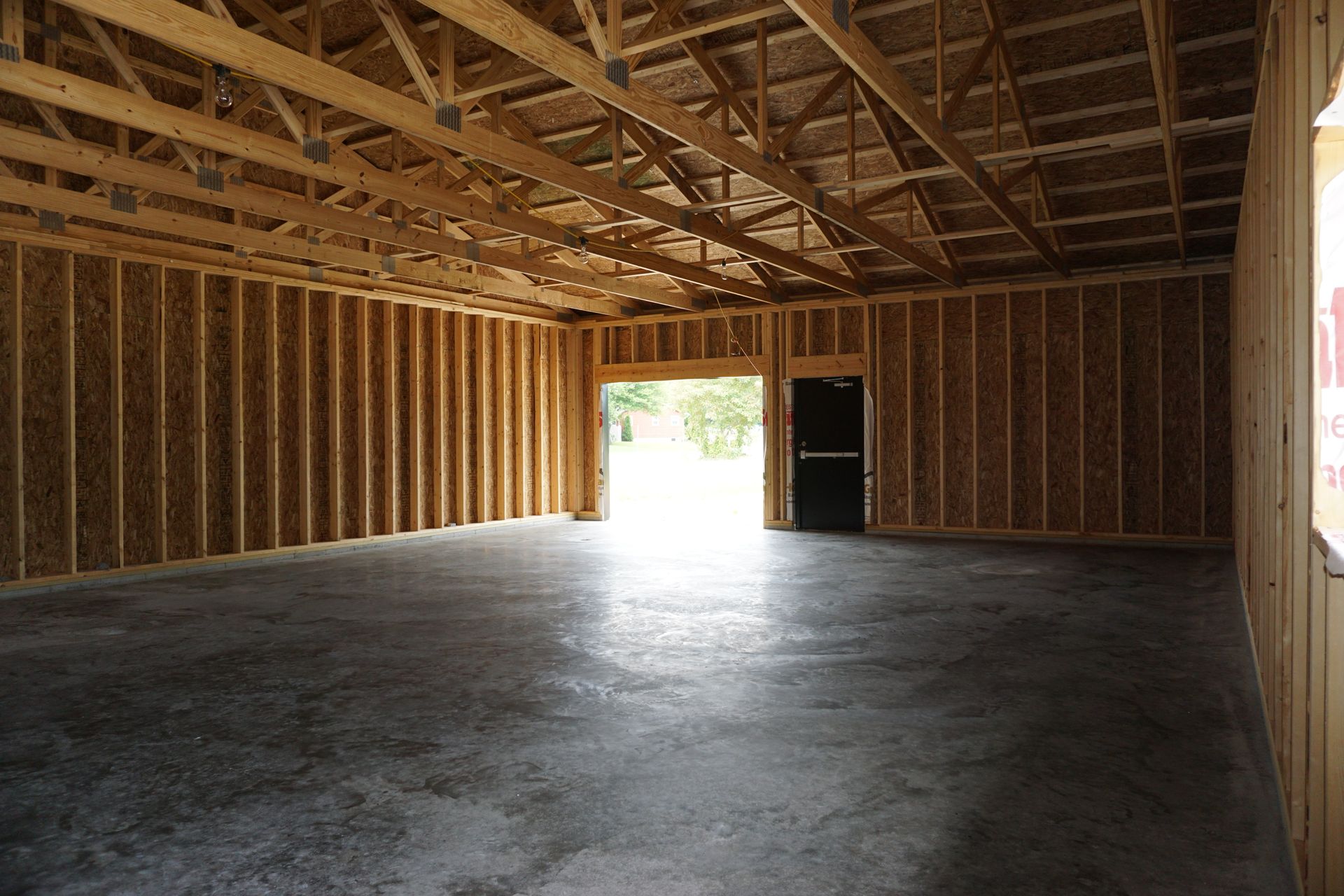 Empty unfinished garage with exposed wooden framing and a concrete floor, lit by daylight from an open doorway.