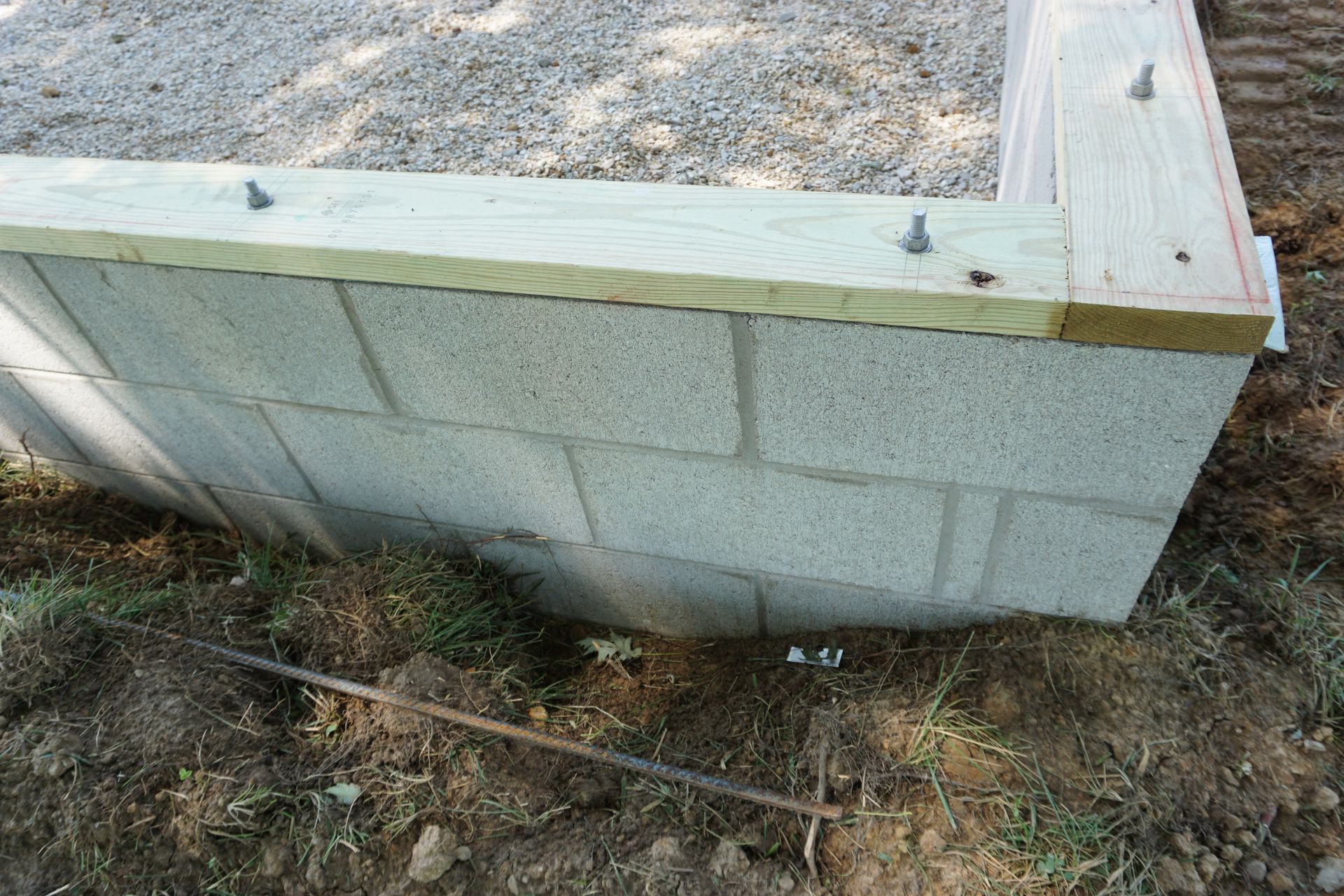 Concrete block retaining wall with wooden cap and bolts, viewed outdoors on gravel and dirt.