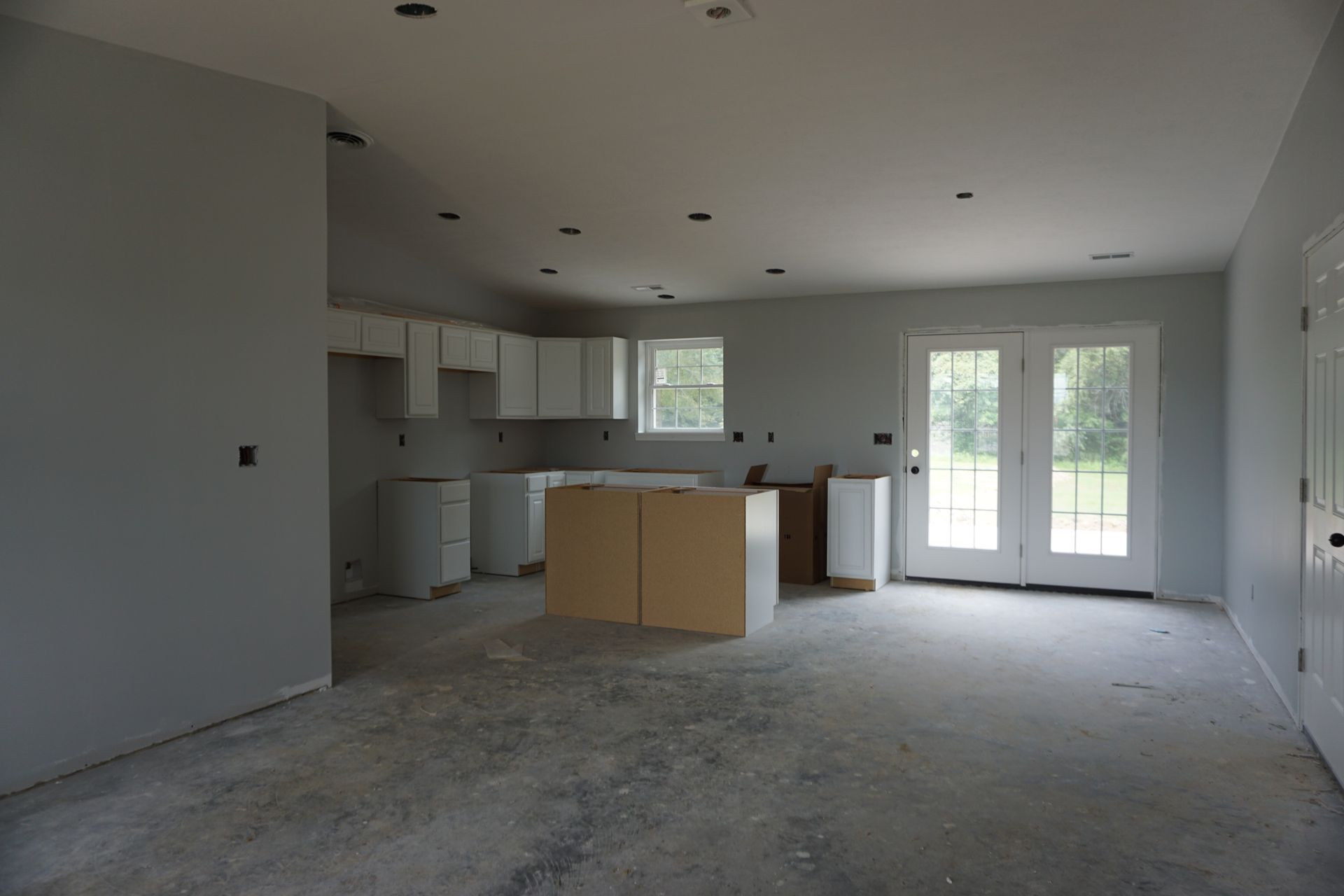 Unfinished modern kitchen and living area with white cabinets, island, and glass doors to outside.