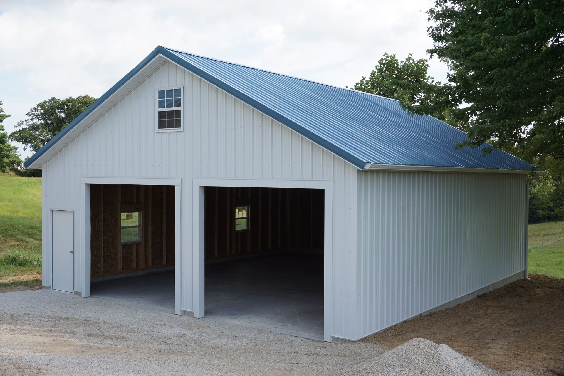 White metal barn with blue roof and two open garage bays on a gravel driveway in a rural setting