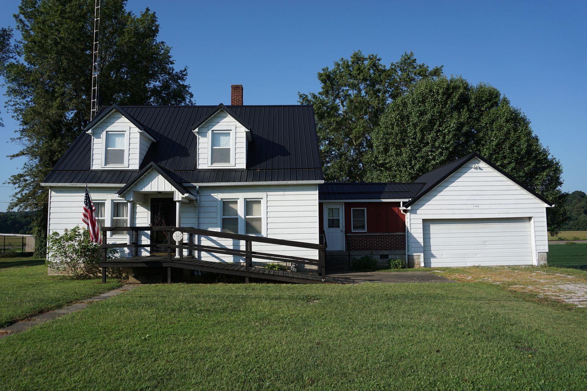 White farmhouse with dark roof, front porch ramp, and attached garage on a grassy lot under a blue sky