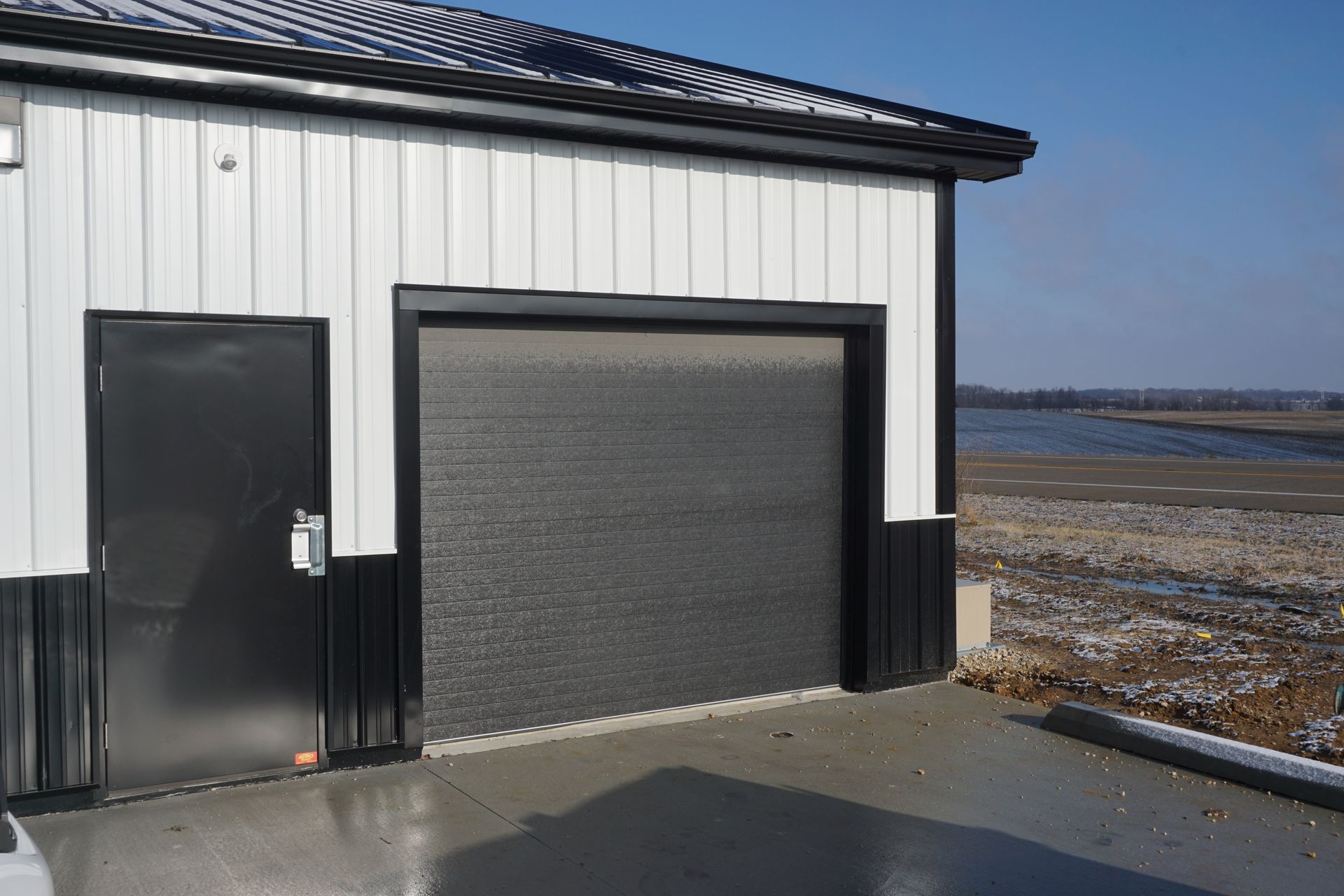 White garage building with a black door and closed roll-up door beside a snowy shoreline