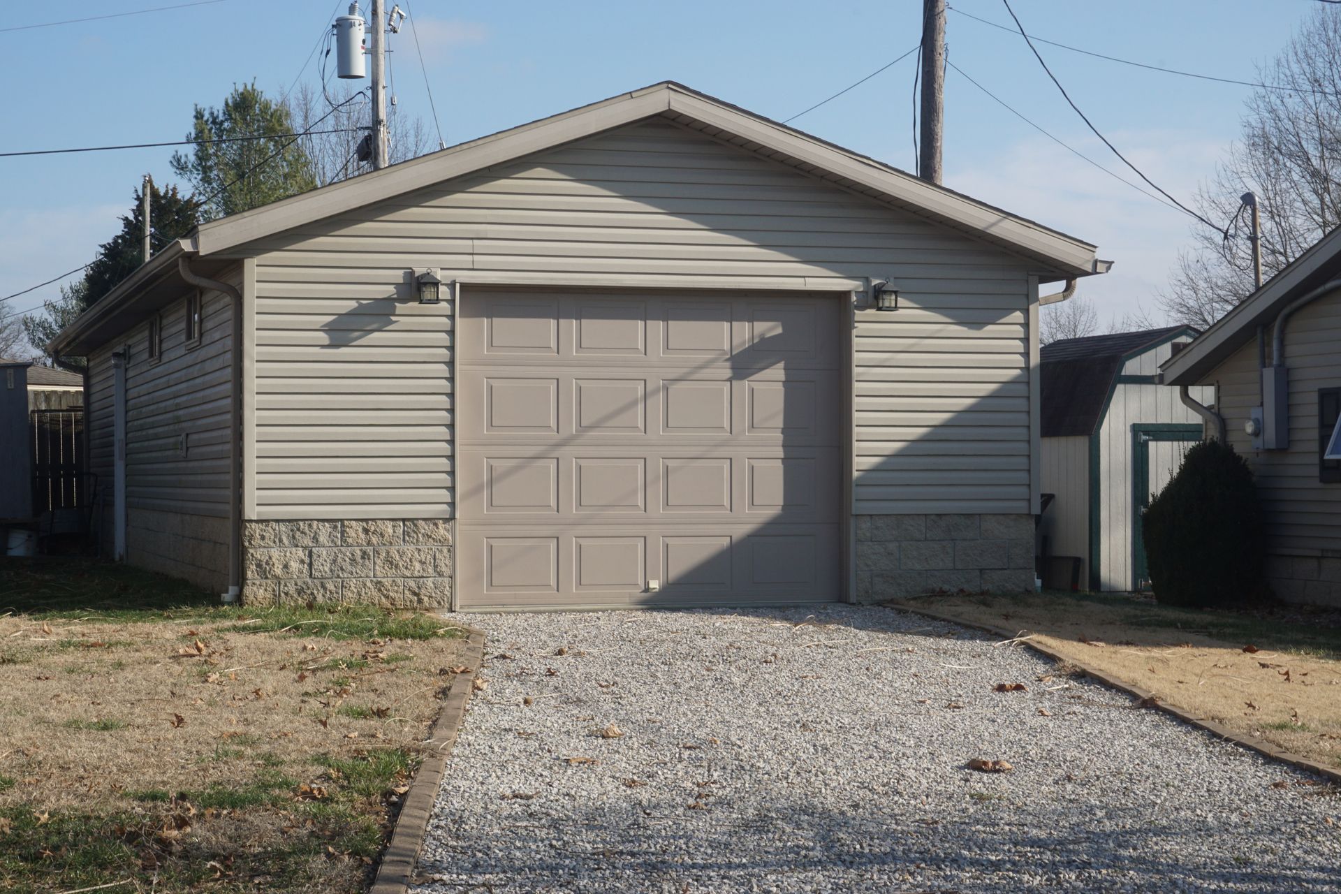 Small beige garage with a closed door and gravel driveway in a residential yard