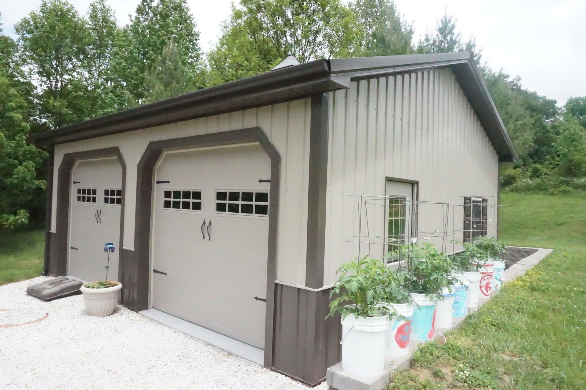 Two-car detached garage with beige siding, brown trim, and flower planters beside a gravel driveway.