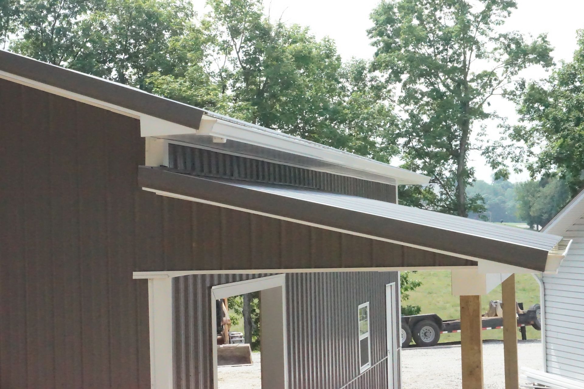 Brown barn with white trim and slanted roof, shown outdoors among trees and gravel.