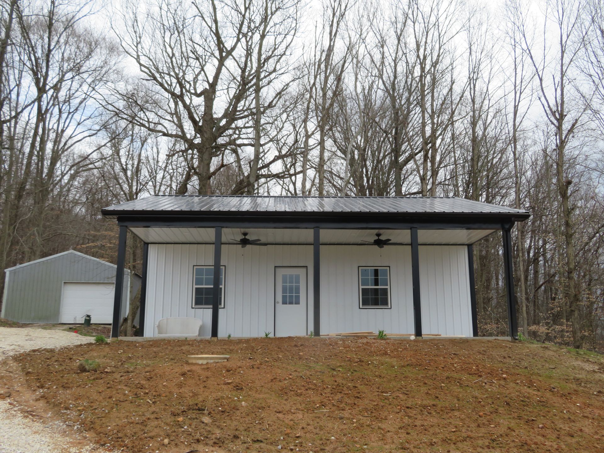 Small white house with a covered porch on a wooded hillside, with bare trees in the background.