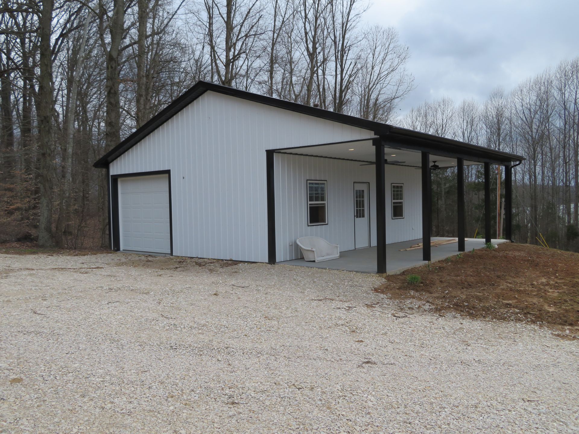 White metal barn with a covered porch beside a gravel lot and wooded background.