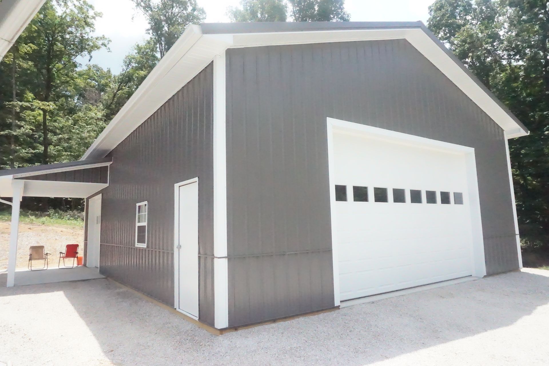 Gray barn-style garage with white trim and a large white roll-up door, set beside trees.
