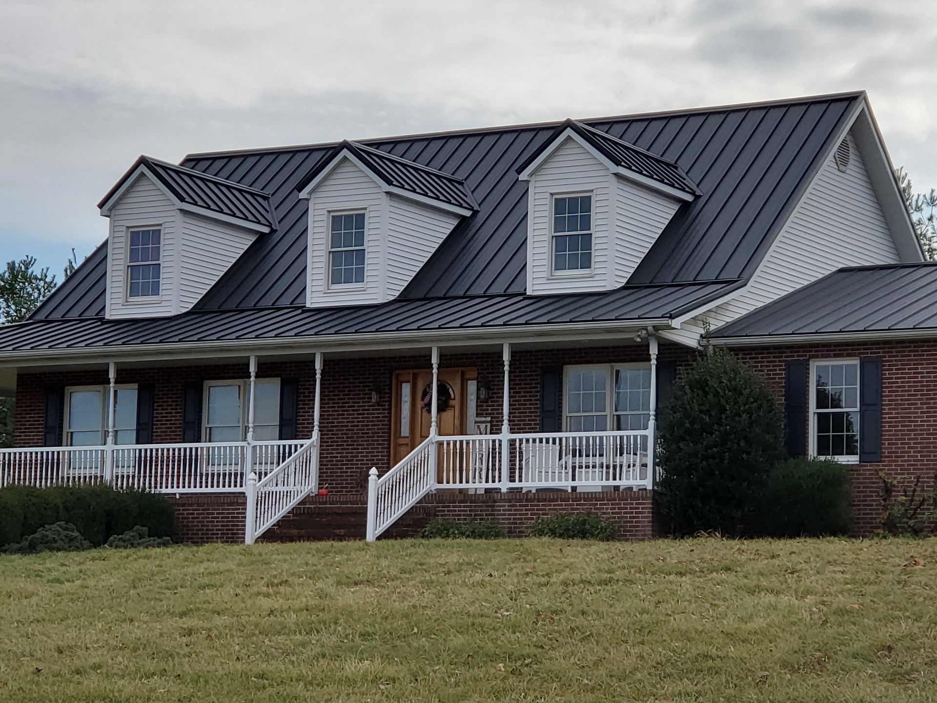 Brick house with dark metal roof, three dormer windows, and white porch railings on a grassy lawn