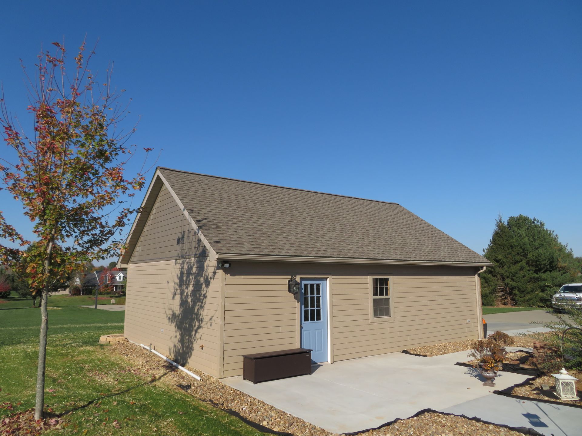 Small tan shed with a blue door and concrete patio in a grassy yard under a clear sky