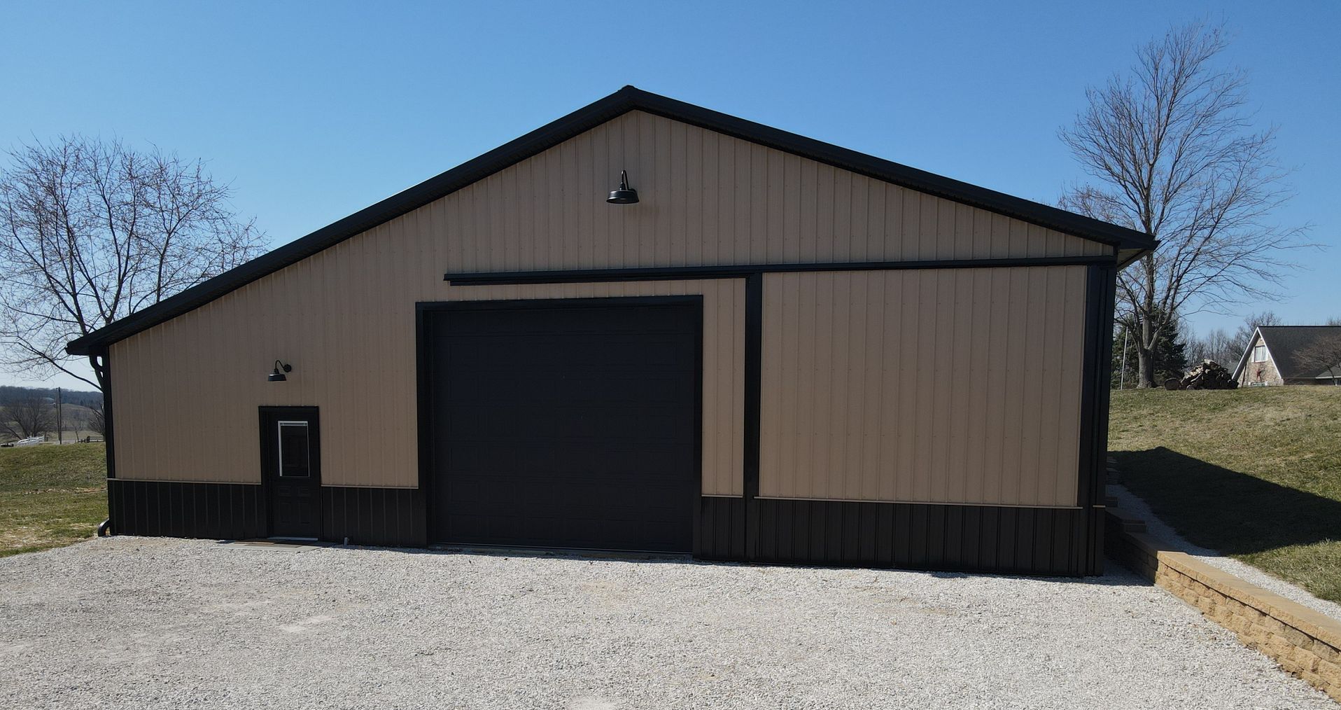 Large beige metal barn with black trim and gravel driveway under a clear blue sky