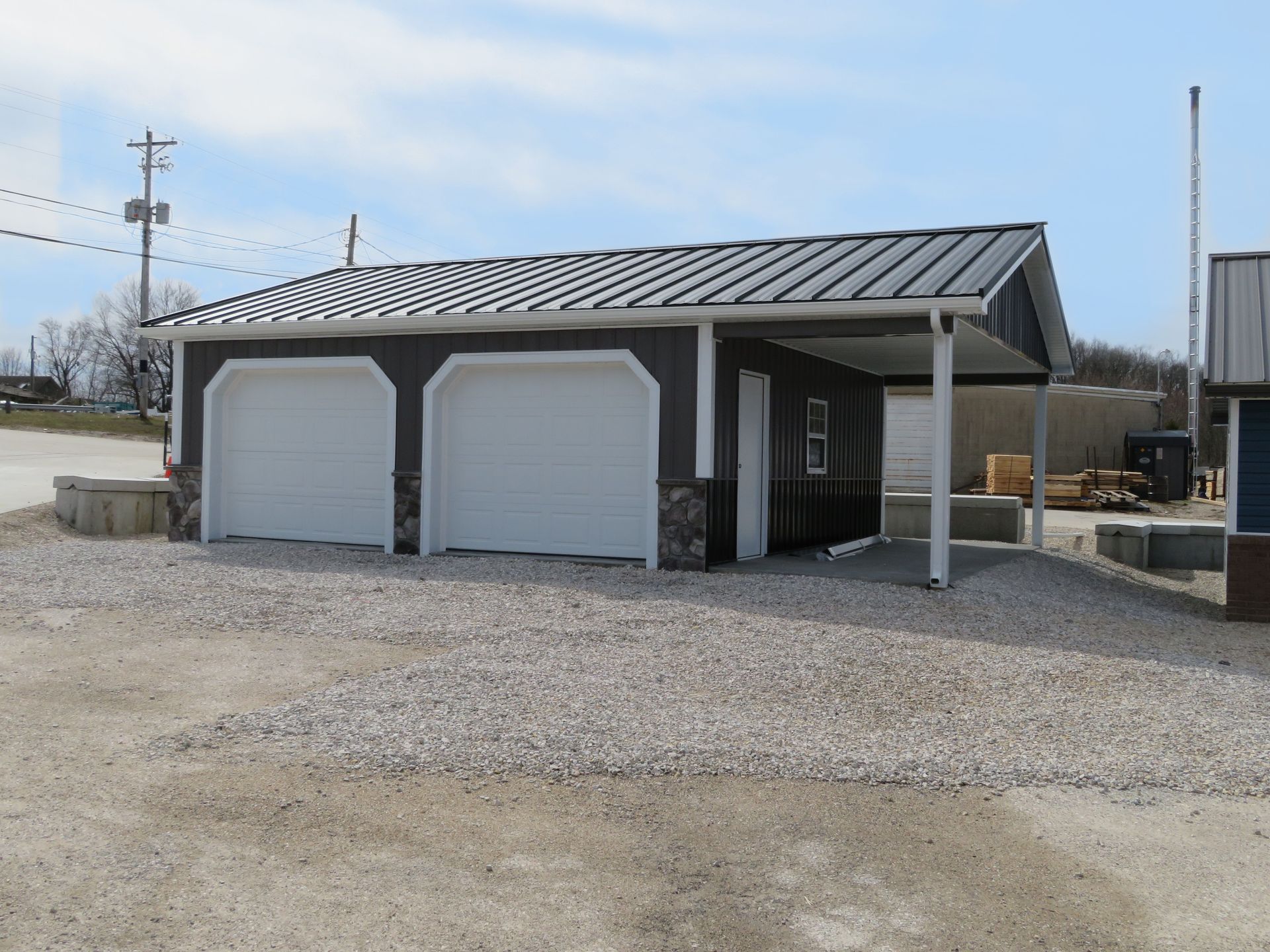 Small gray two-bay garage with white doors on a gravel lot under a blue sky