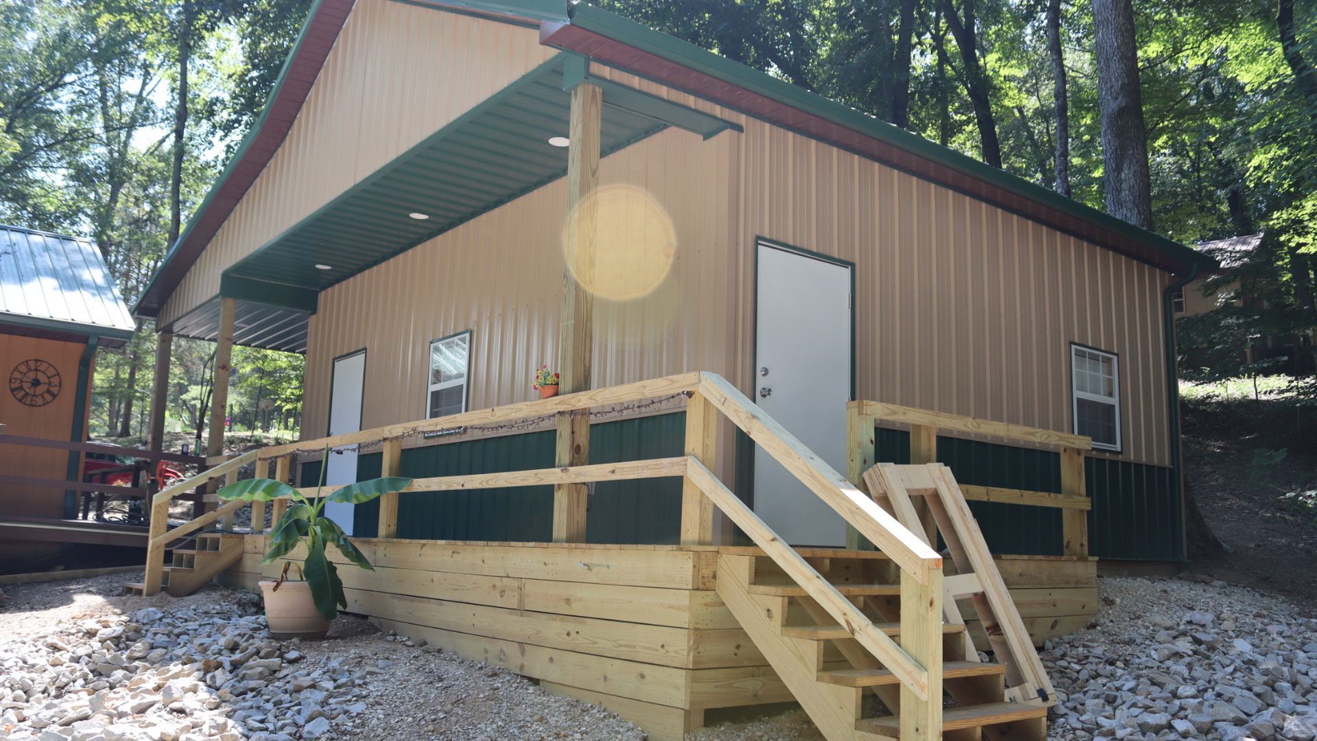Small beige cabin with a wooden ramp and stairs in a wooded setting, sunlight filtering through trees