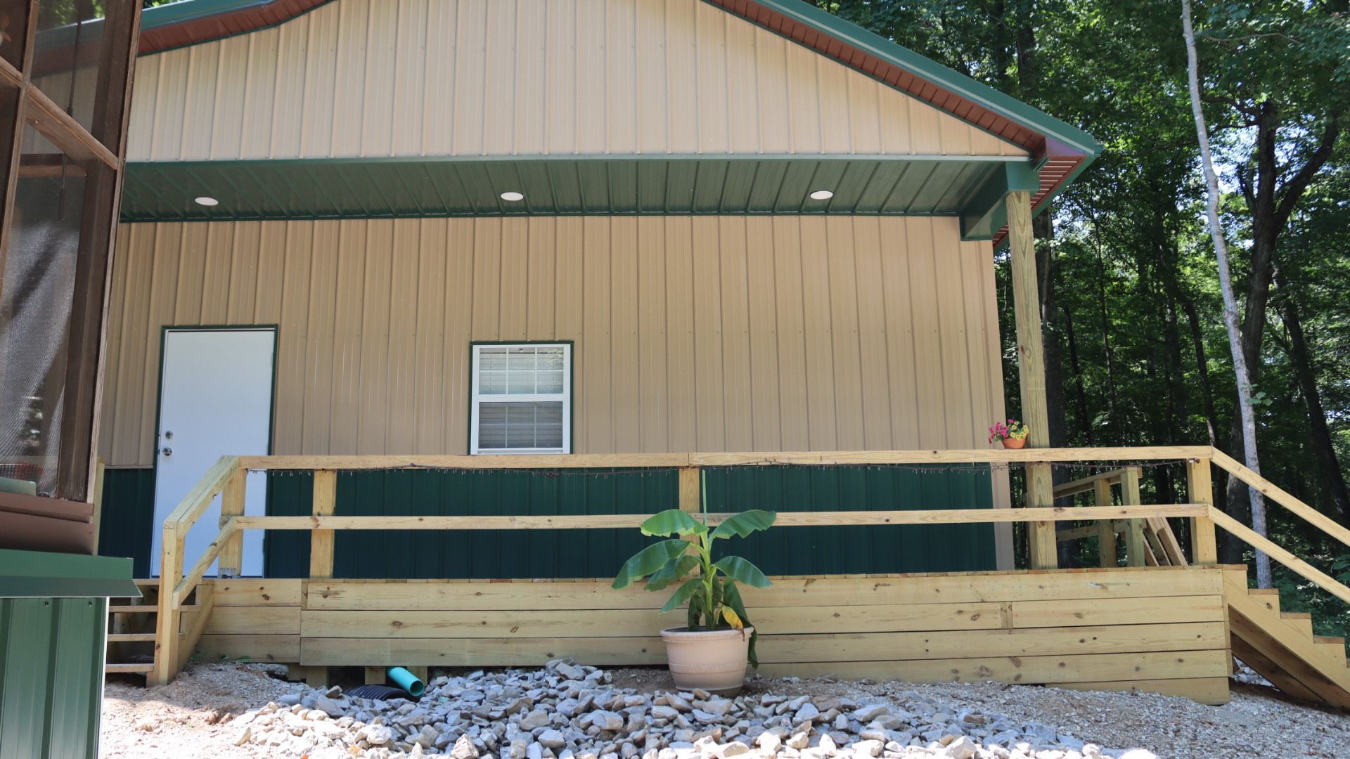 Tan house exterior with a covered porch, wooden ramp railing, and potted plant on a gravel yard.