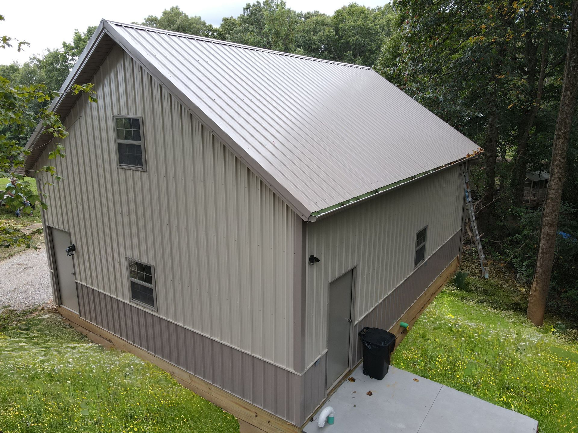 Gray metal-sided barn with a steep silver roof on a grassy hillside beside trees