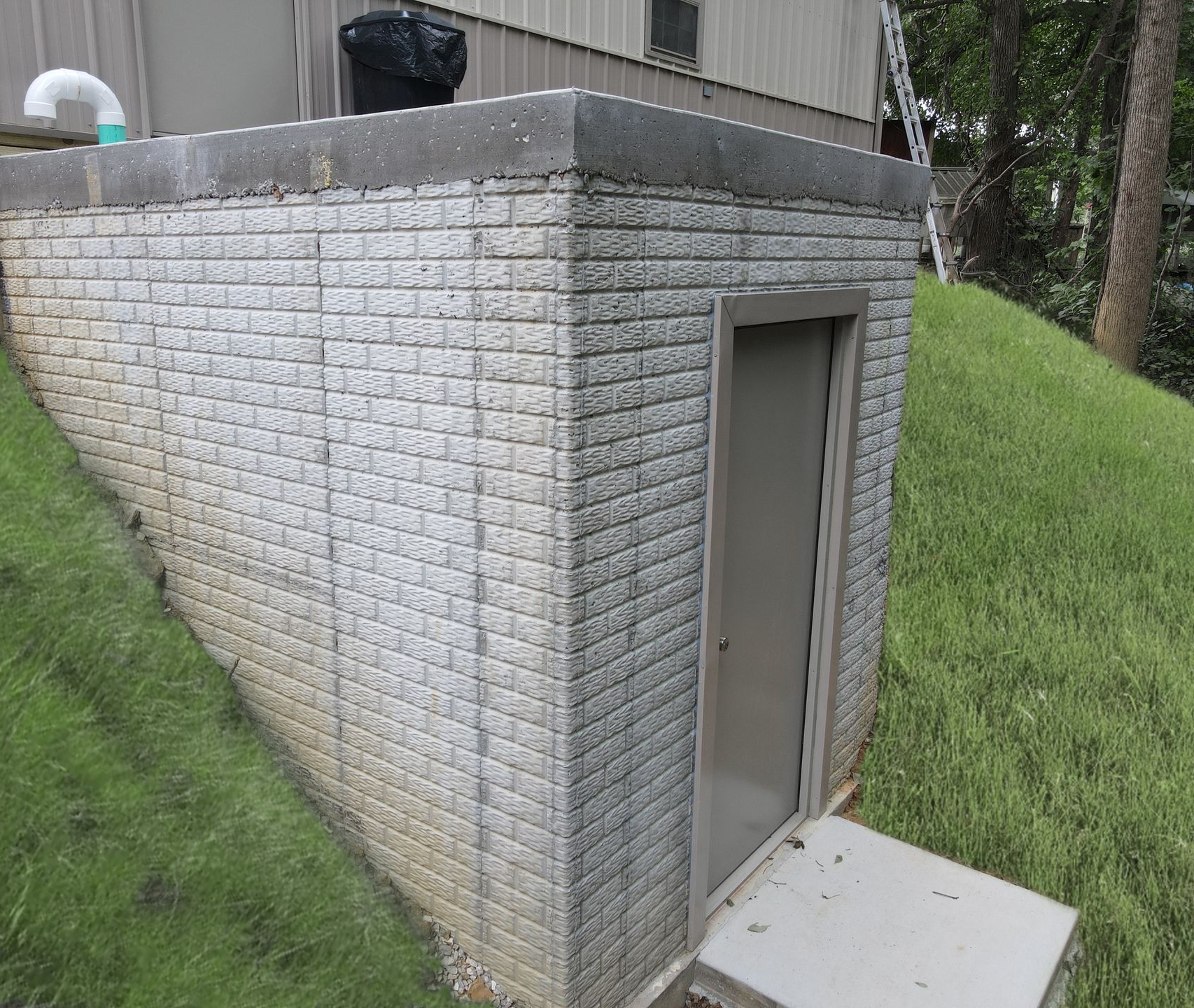 Small concrete utility building with a gray door beside a grassy slope and trees.