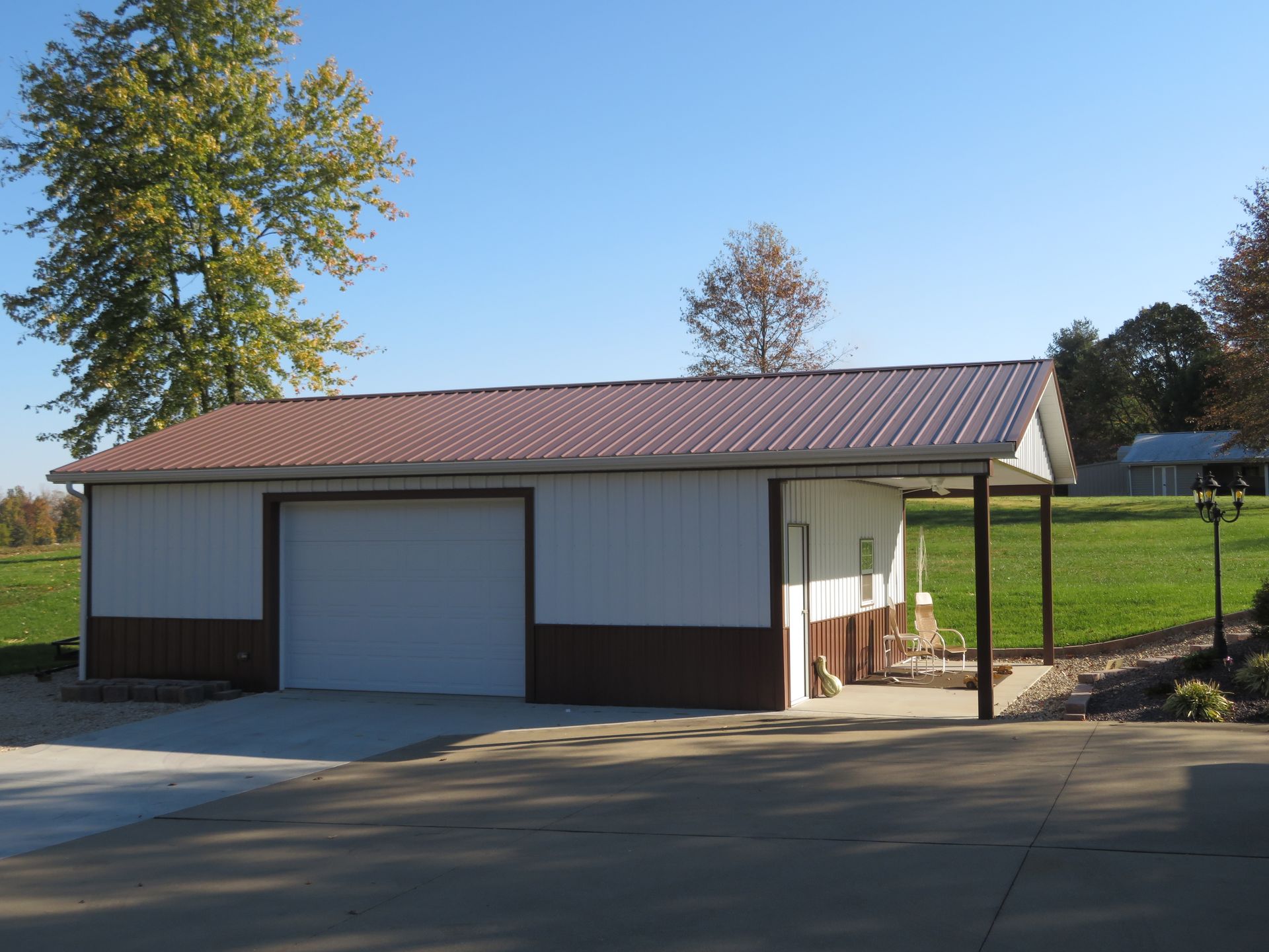 Small white garage with brown trim and red roof beside a driveway on a sunny rural property