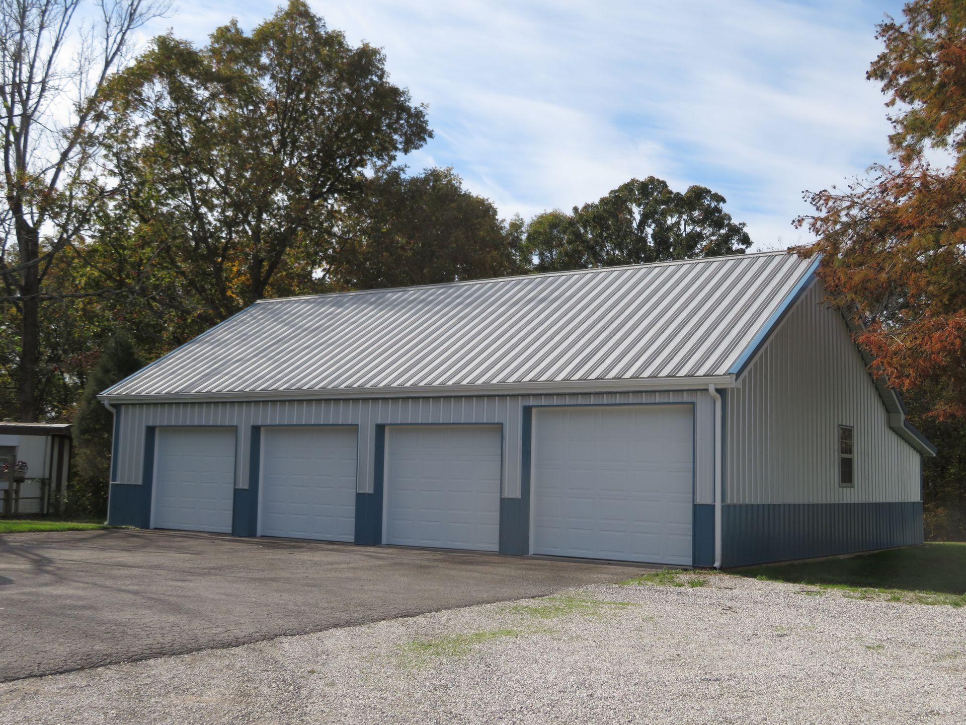 Gray metal garage with four doors on a gravel lot, surrounded by trees under a partly cloudy sky