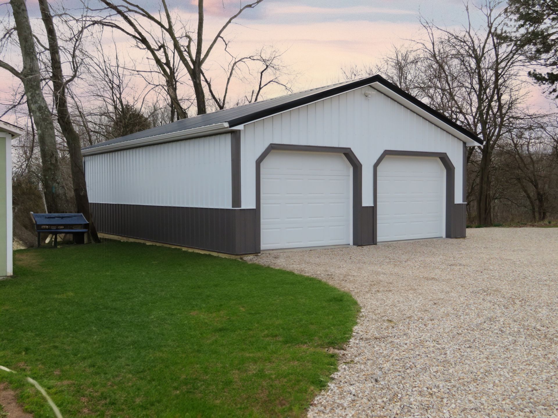 White two-car garage with dark trim beside a gravel driveway and green lawn at dusk