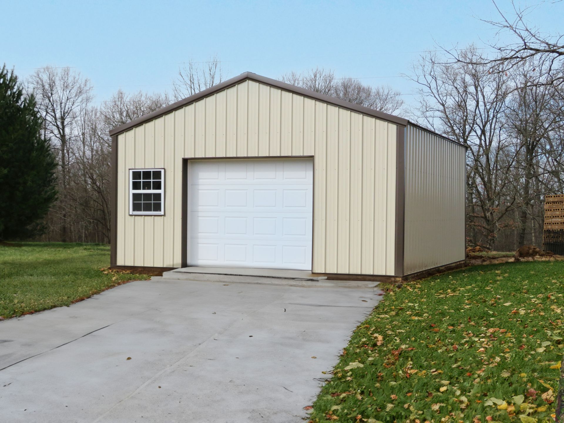 Beige metal garage with white door and driveway in a wooded yard