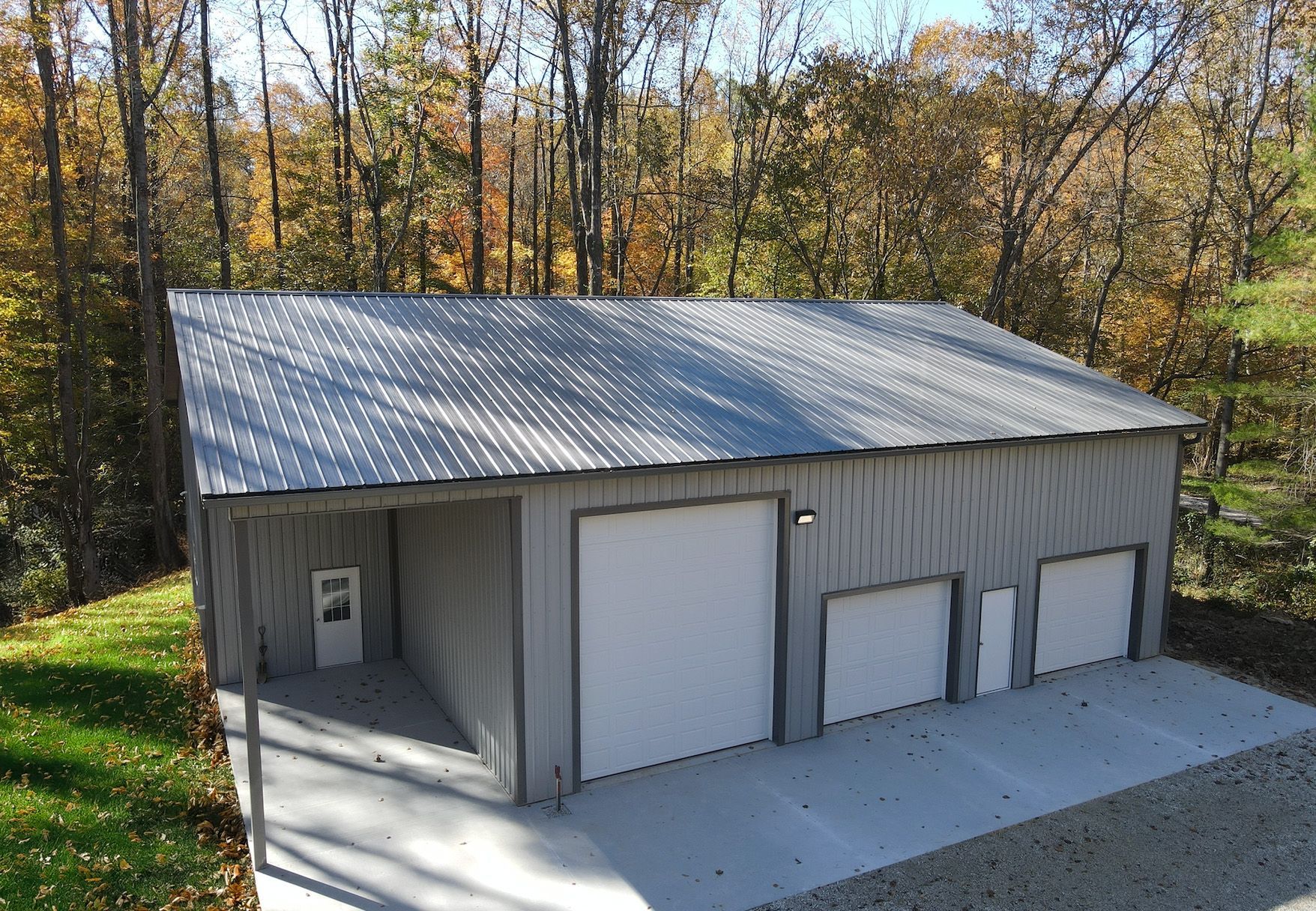 Small gray metal garage with three white doors beside a wooded hillside in autumn