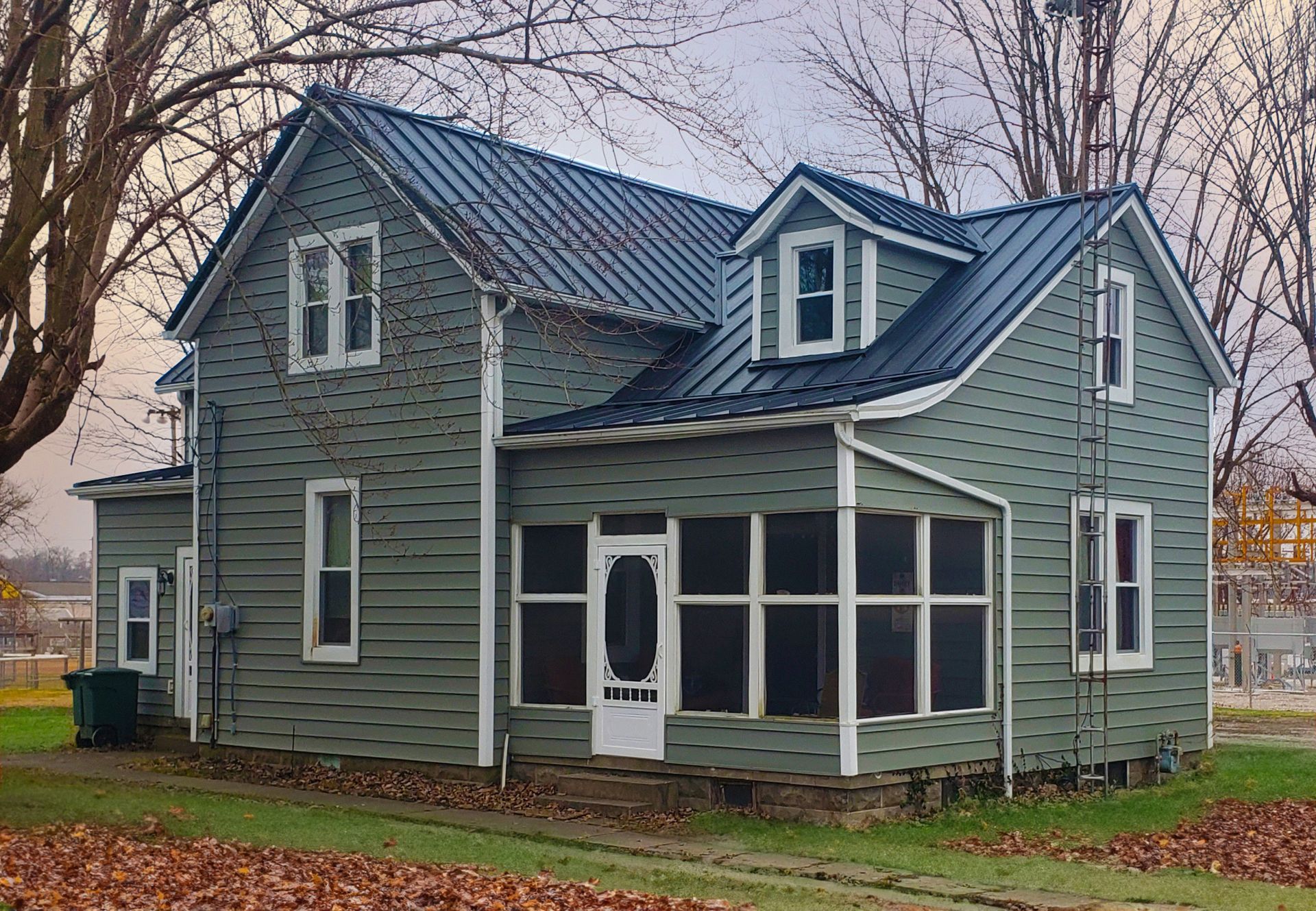 Green two-story house with a screened front porch and dormer windows in a leaf-covered yard.