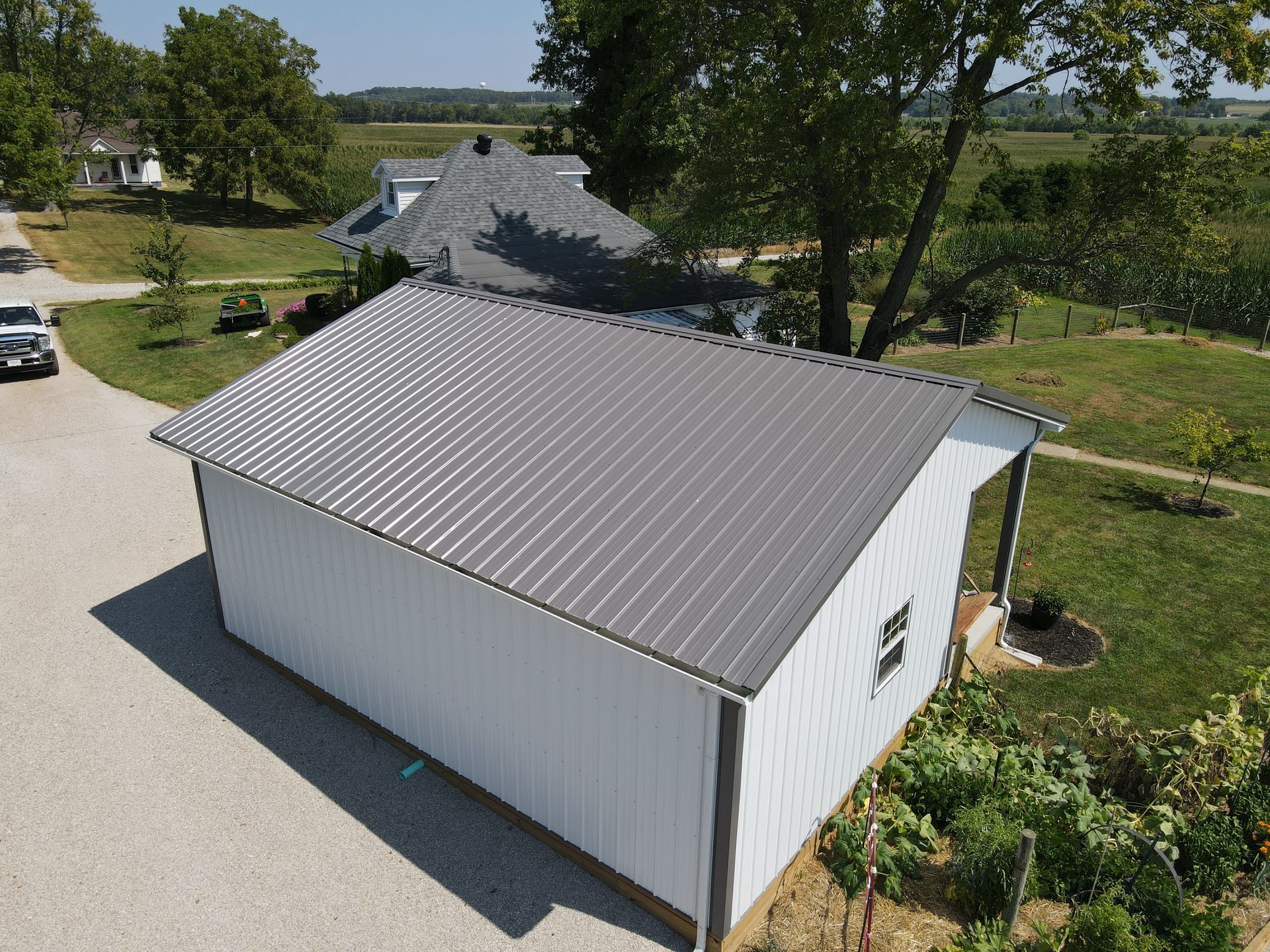White metal-roofed outbuilding beside a garden and trees on a rural property