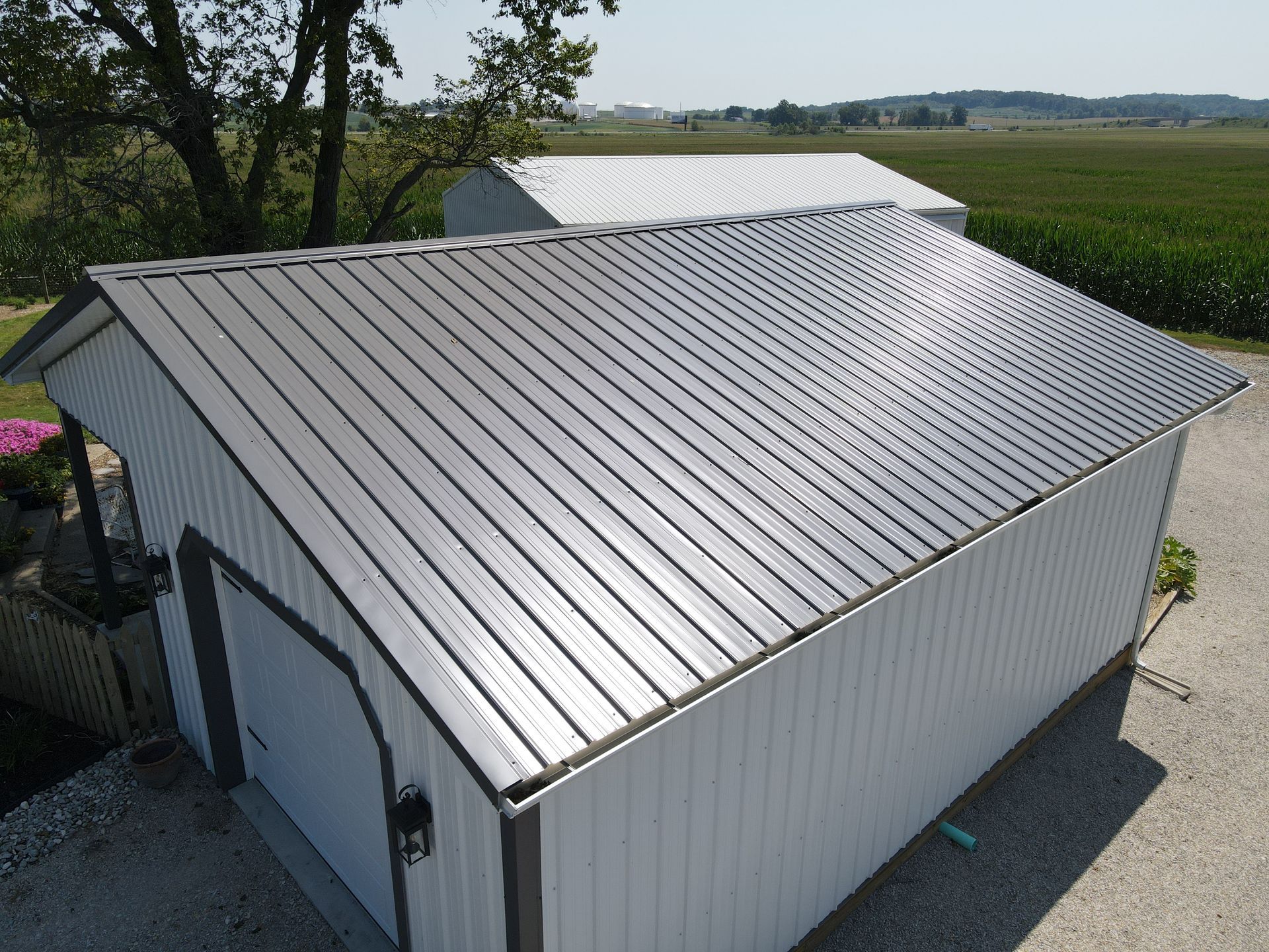 White metal-roofed shed beside a gravel drive, with fields and trees in the background.