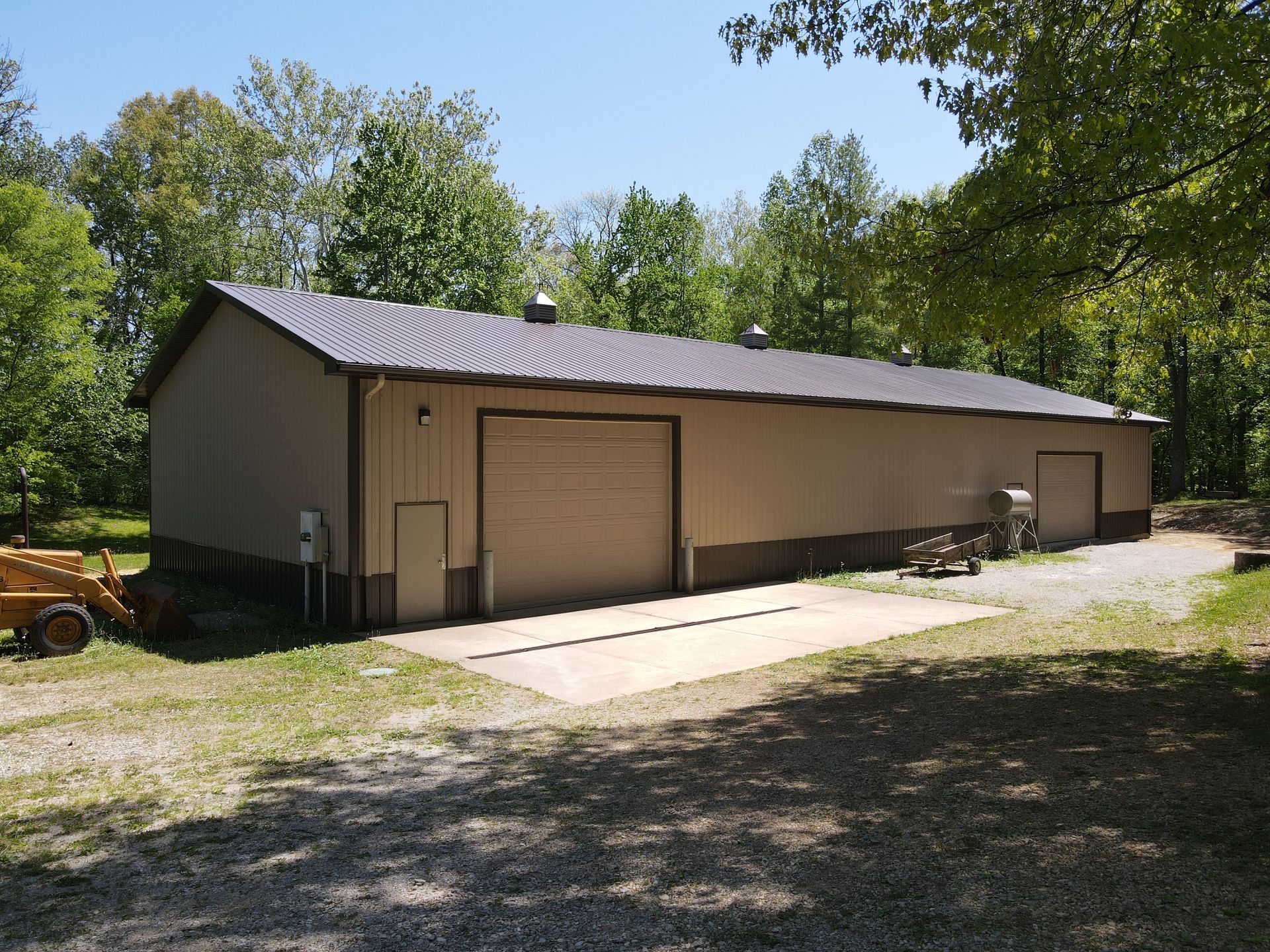Long tan metal garage with two roll-up doors beside a gravel driveway, surrounded by trees.