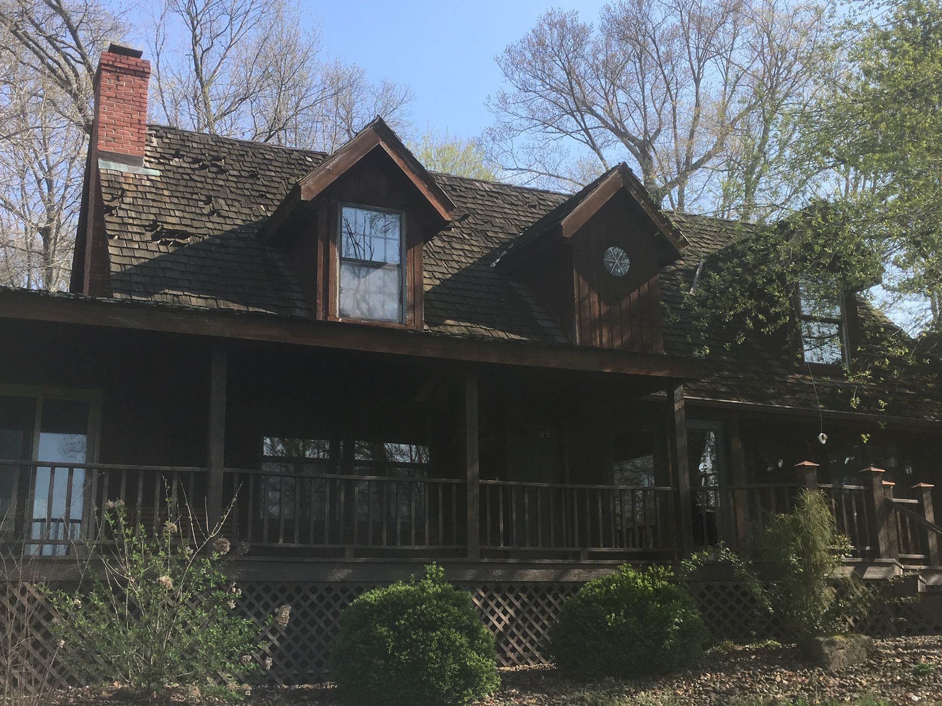 Old dark wooden house with a porch, steep roof, and brick chimney amid leafless trees.