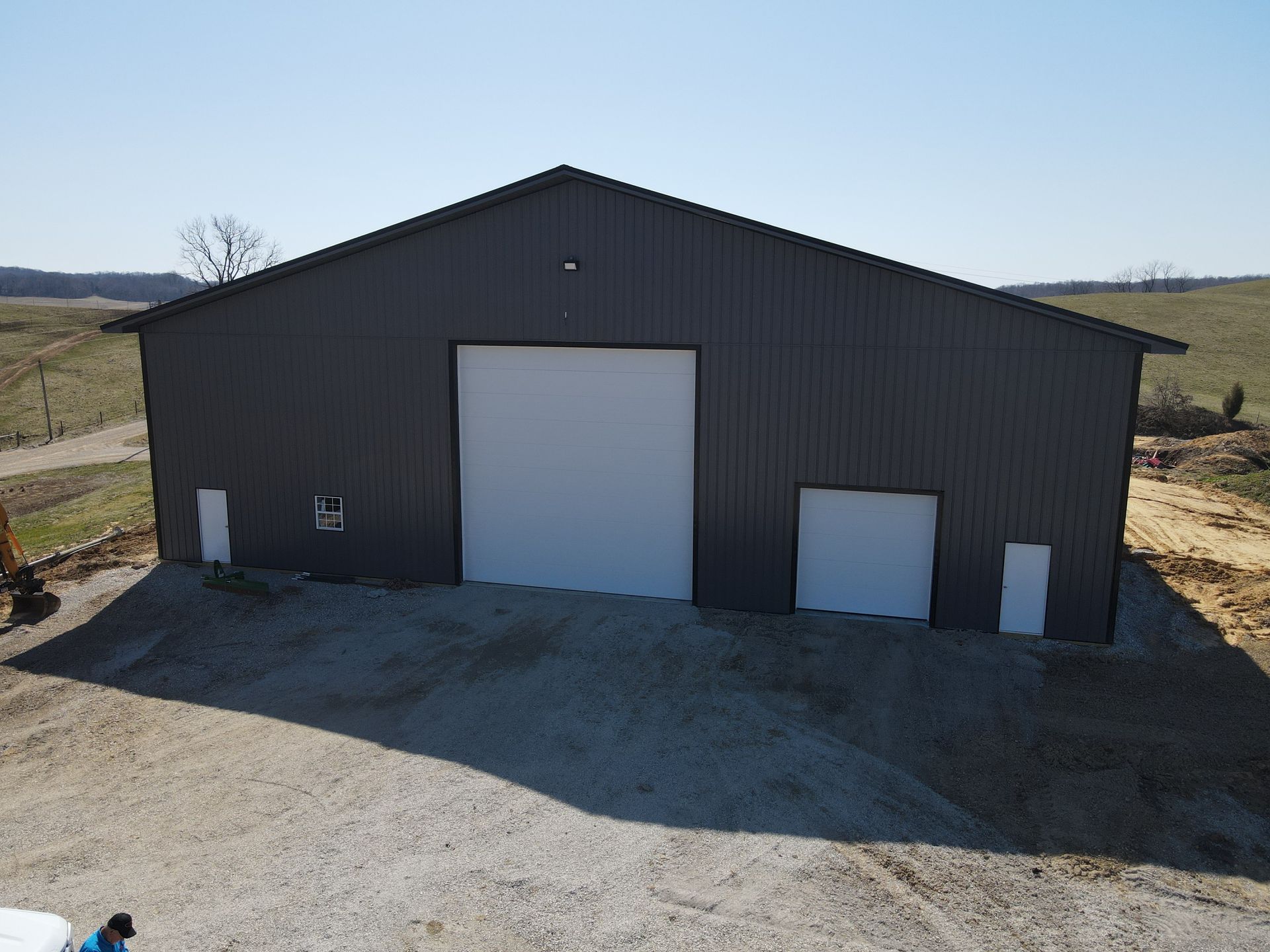 Large dark barn with white doors on a gravel lot in a rural field, under clear daylight