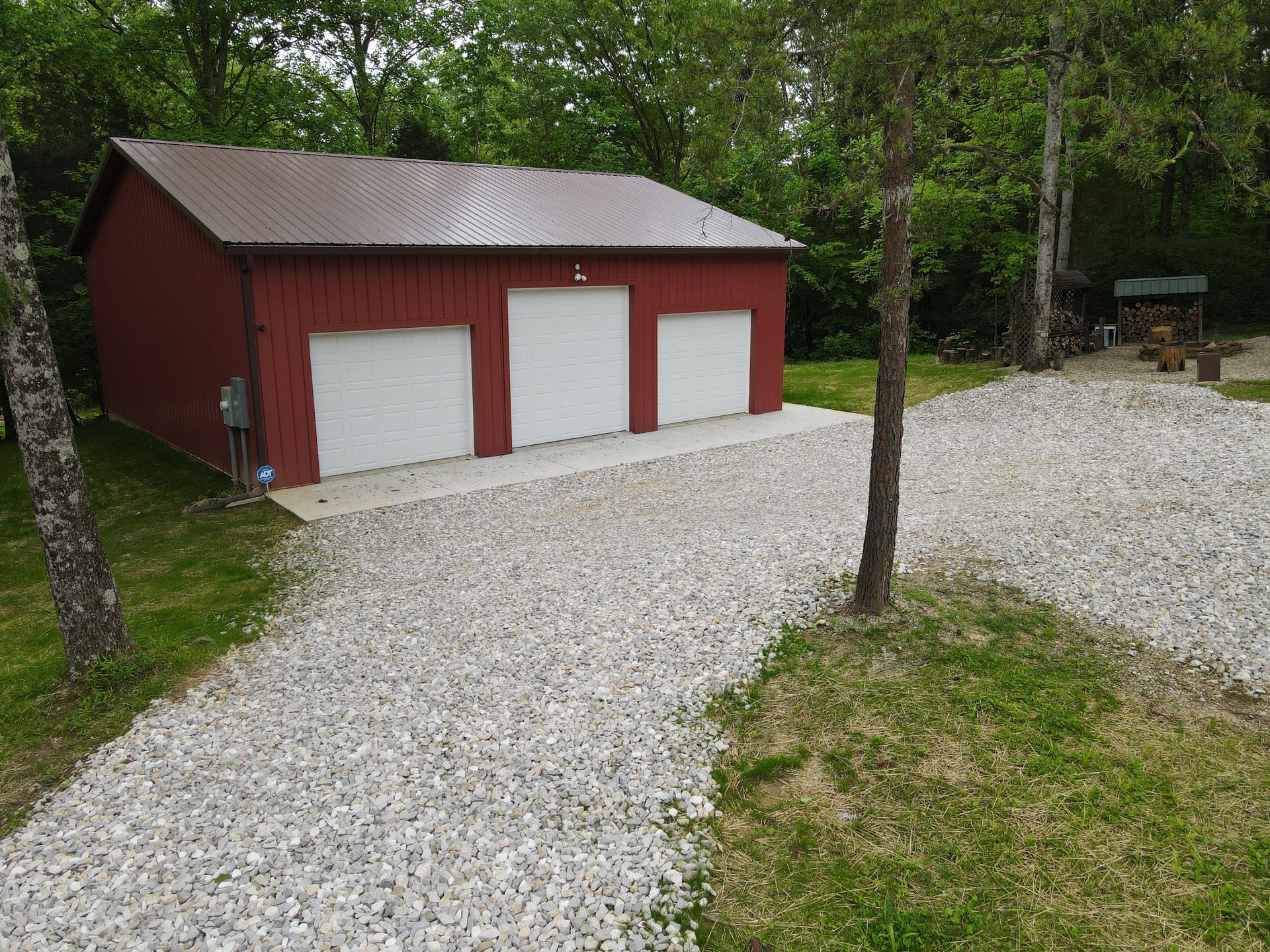 Red three-bay garage beside a gravel driveway, surrounded by trees and greenery.