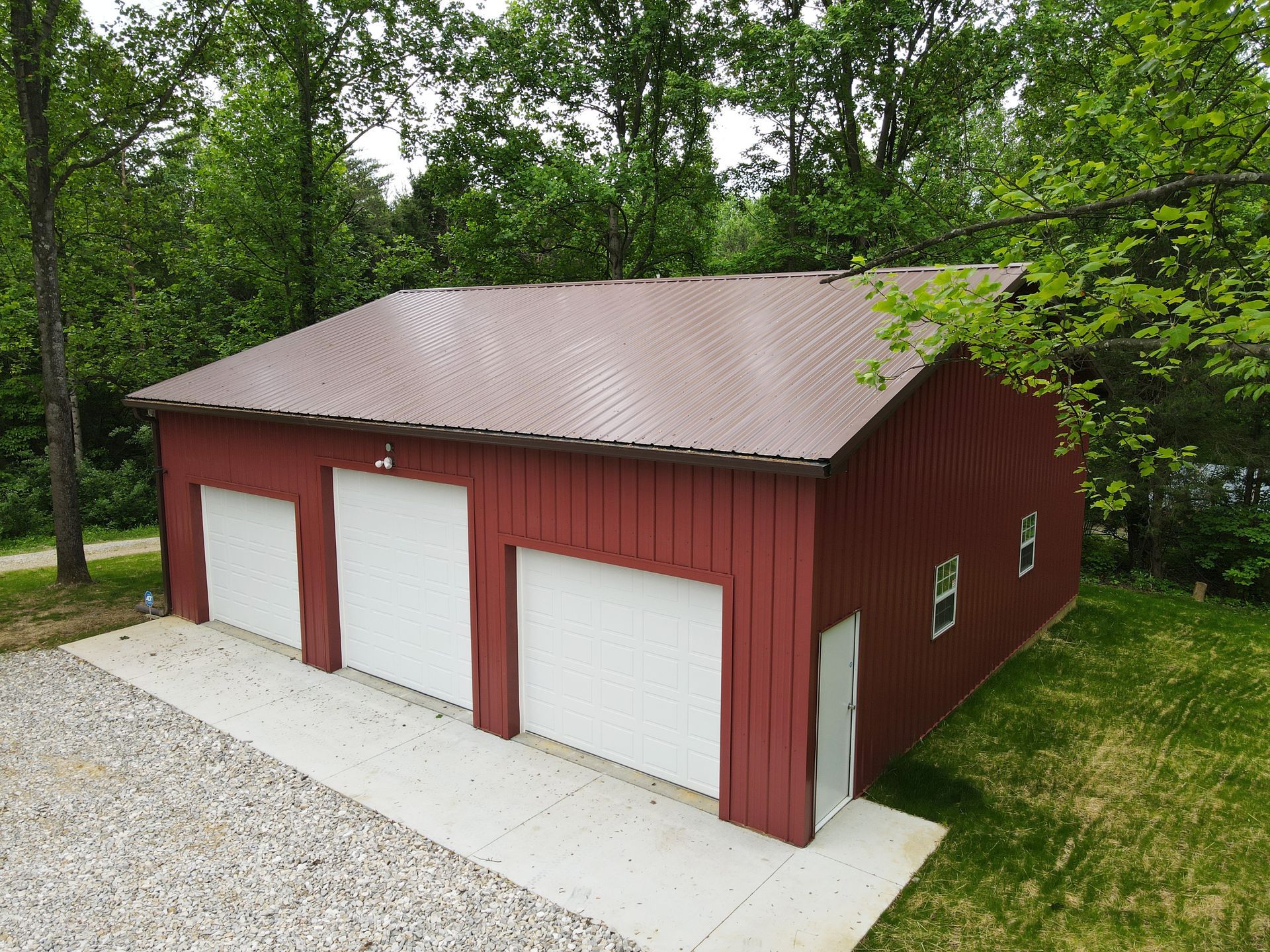 Red barn-style garage with three white doors, brown roof, set in a wooded yard