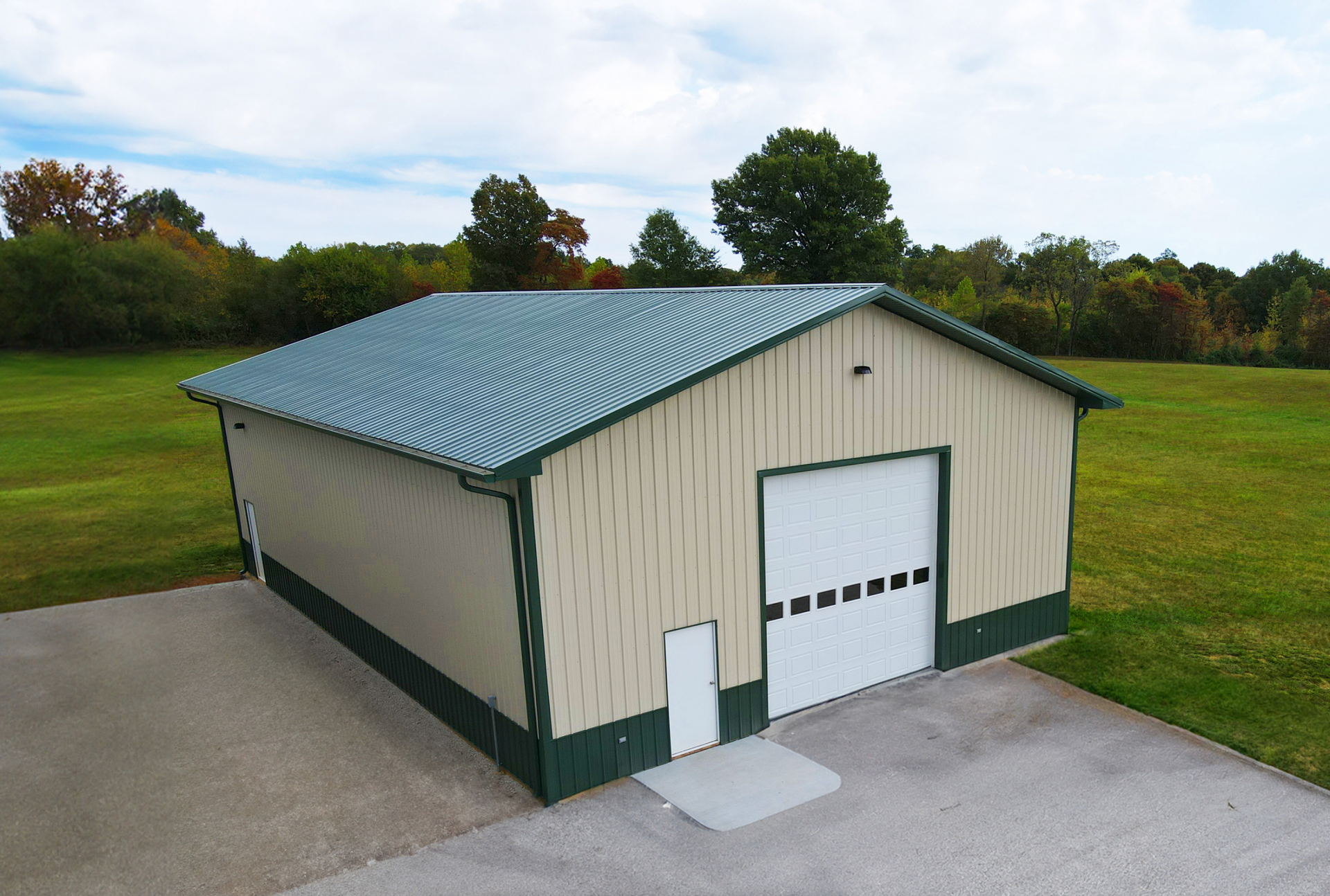 Beige metal garage building with green trim and a green roof on a paved lot, surrounded by grass and trees