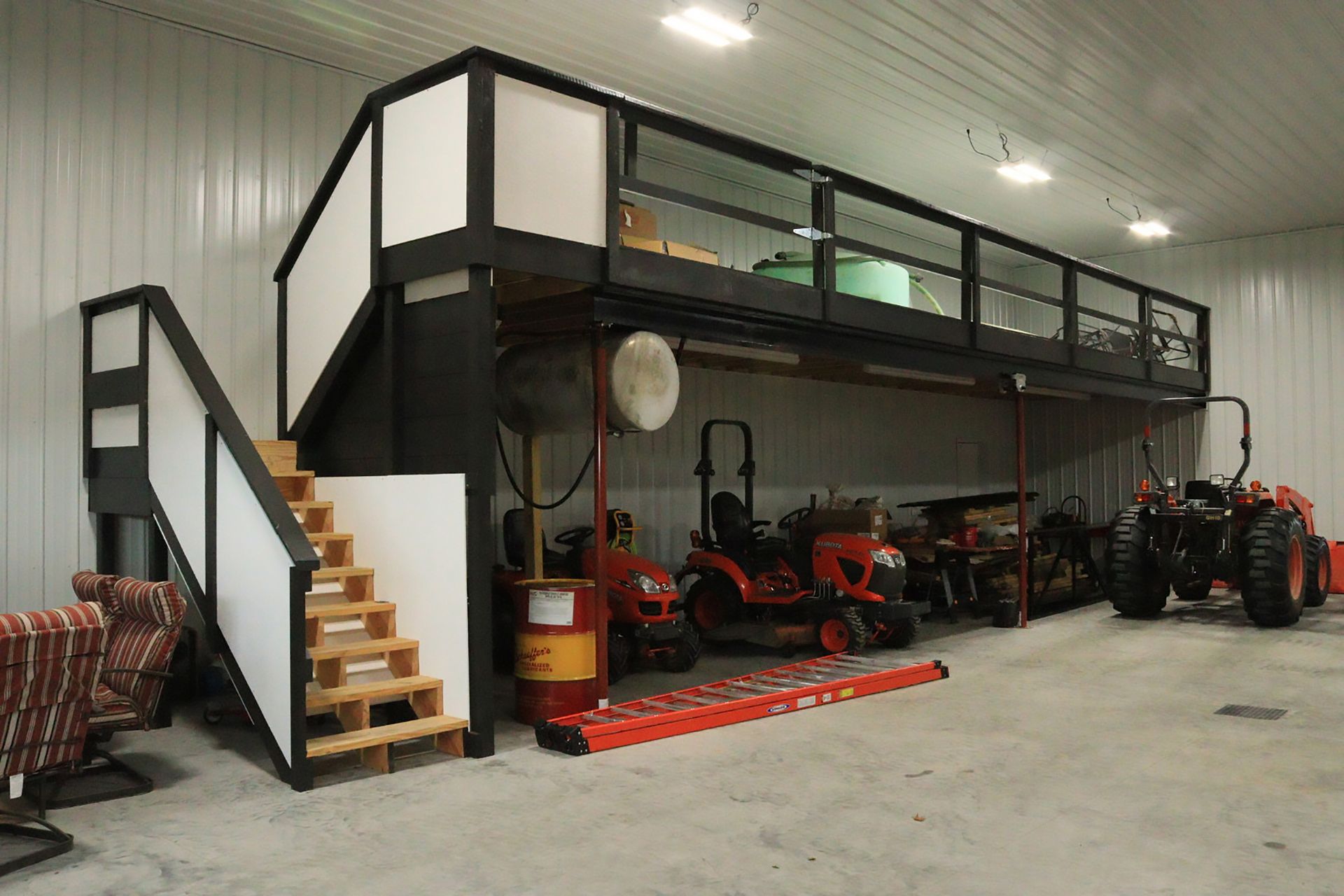 Lofted workshop with stairs, tractors, tools, and a red floor mat inside a large metal garage