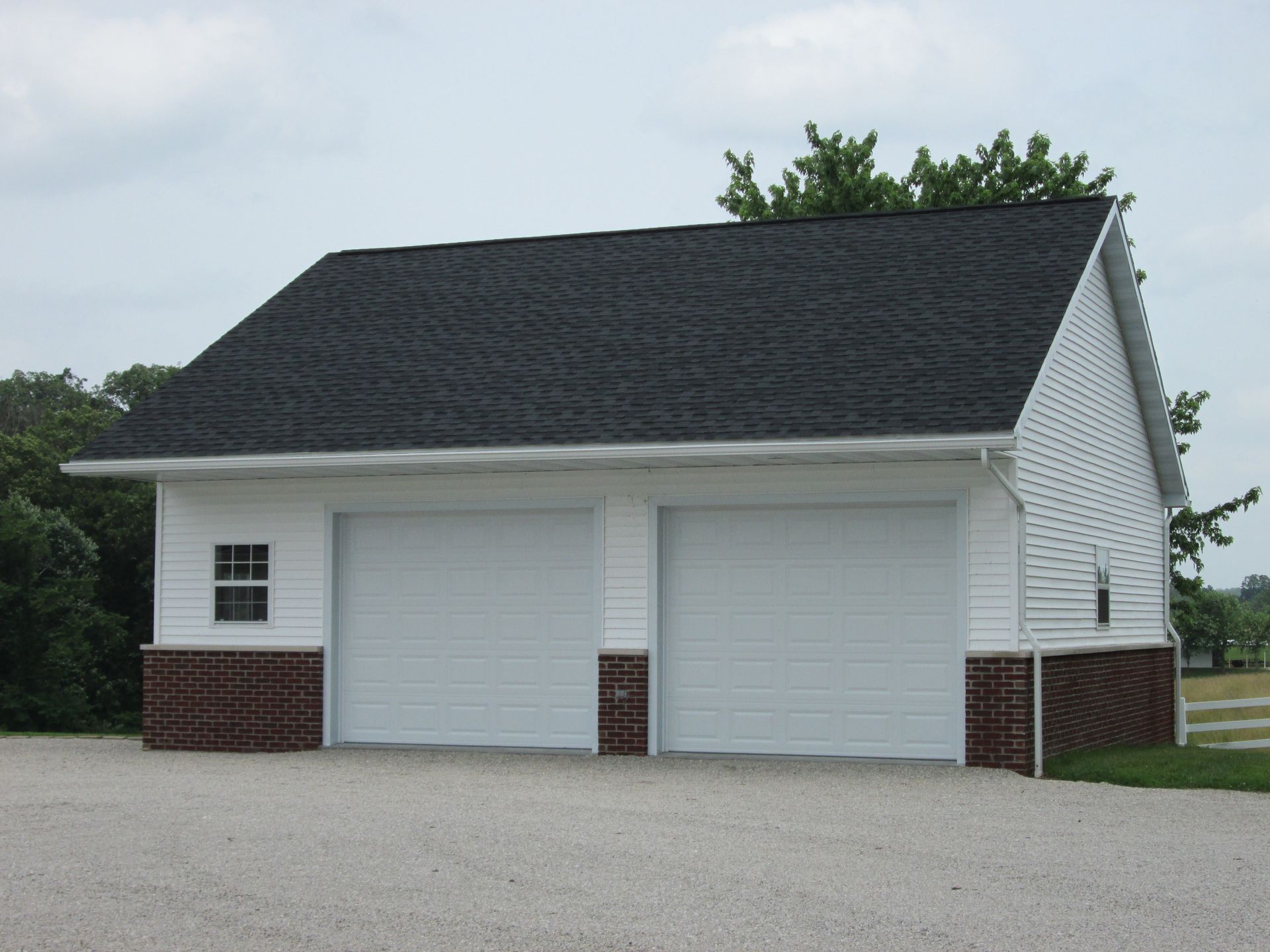 Detached two-car garage with white siding, brick base, and dark shingled roof on a gravel driveway